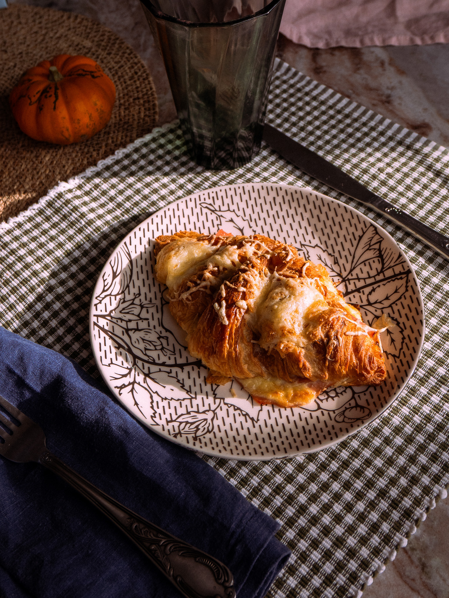 A croissant-style pastry sits on a decorative plate alongside silverware and a small glass, with a mini pumpkin in the background.