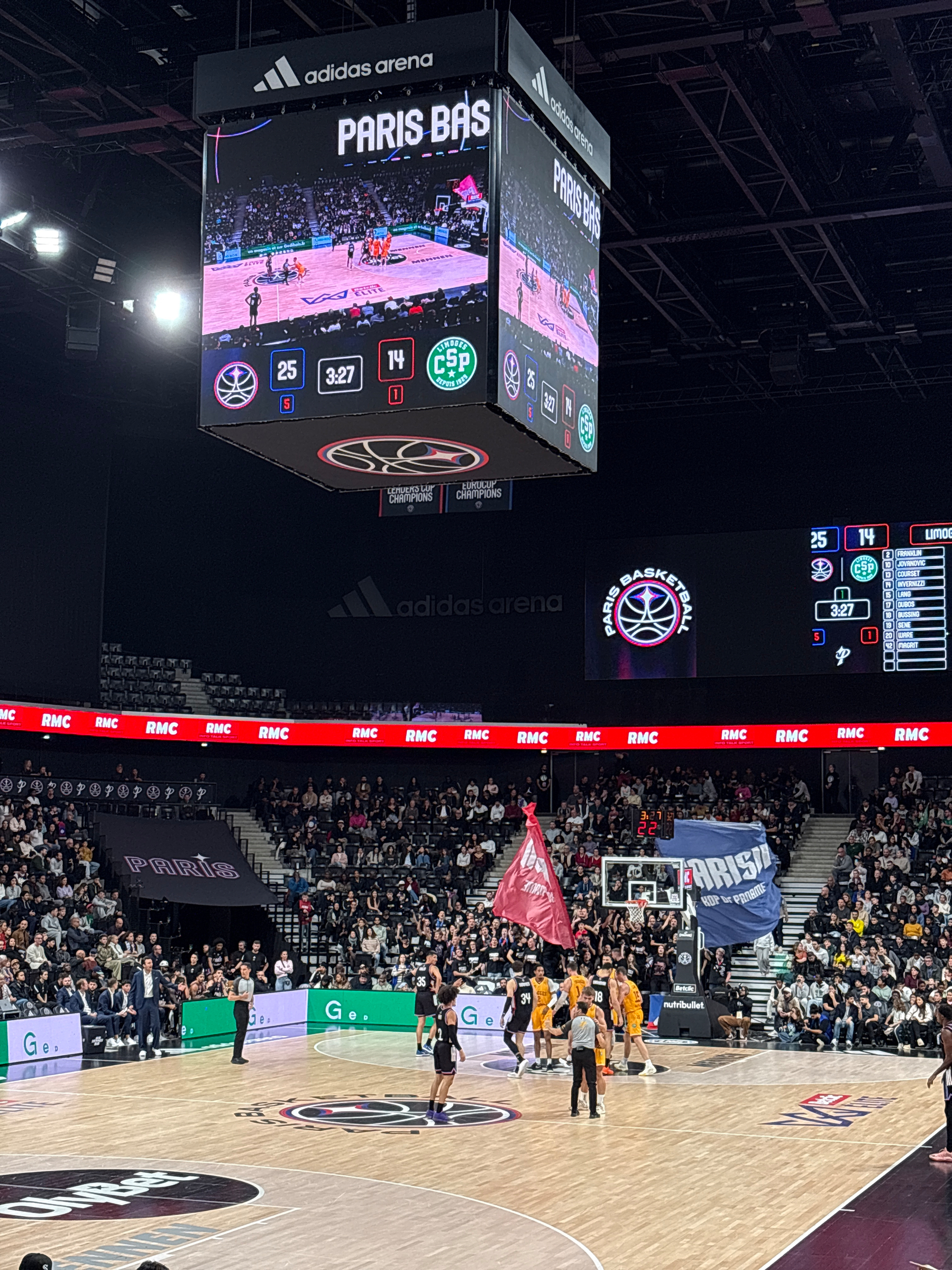 A basketball game is taking place in a large indoor arena with a scoreboard and screen displaying the action above the court.