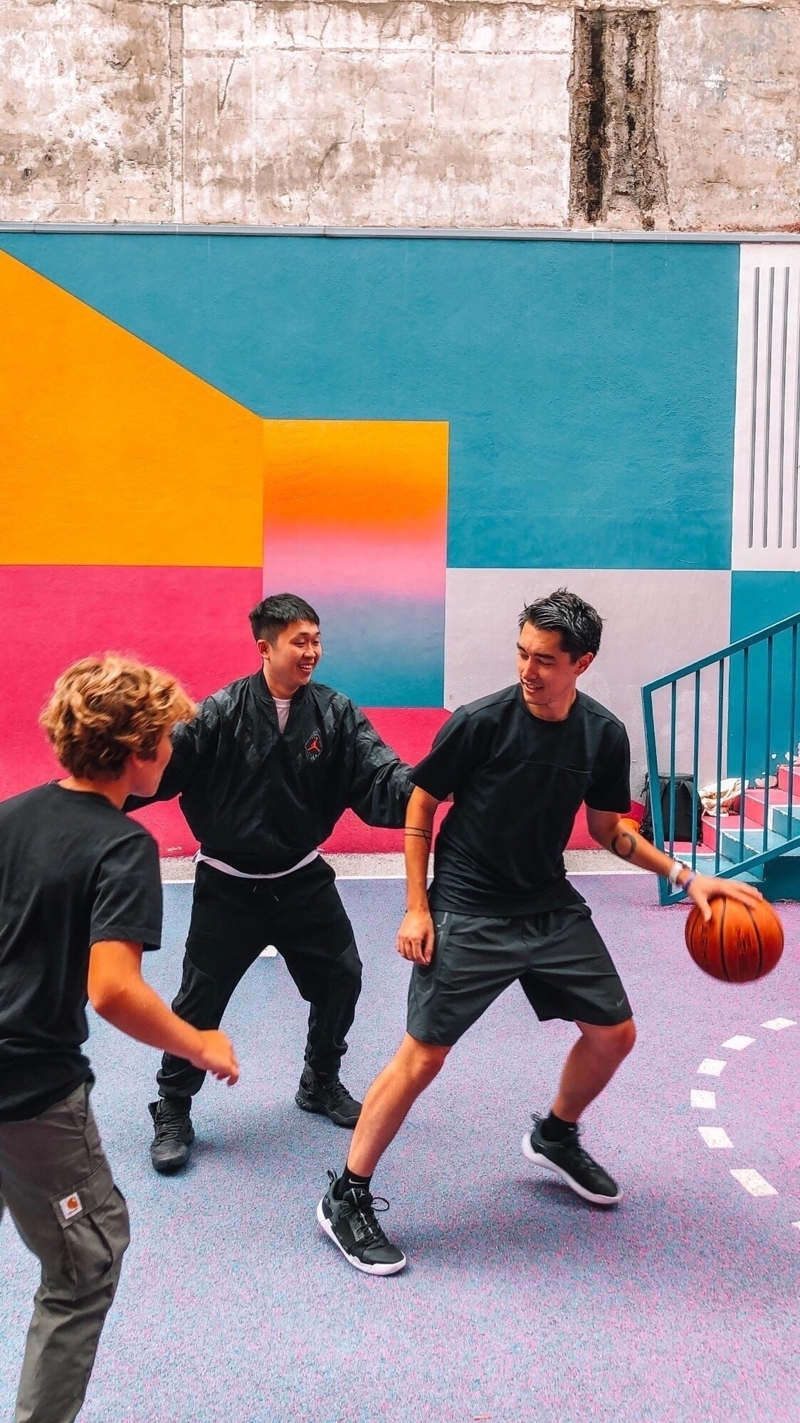 Two people are playing basketball on a colorful court while a third person watches and laughs.