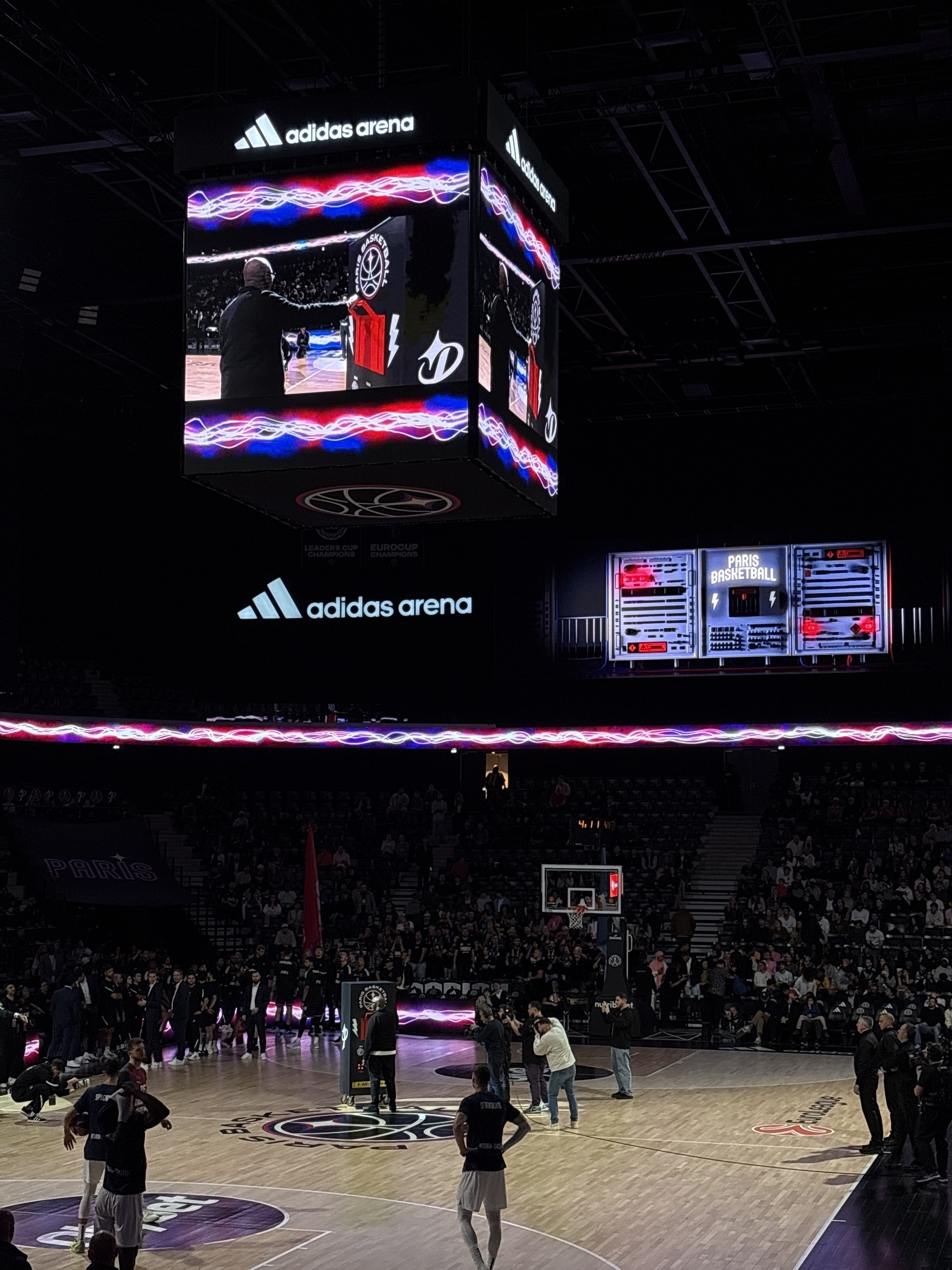 A basketball game is taking place in a large indoor arena with a digital scoreboard and spectators around.