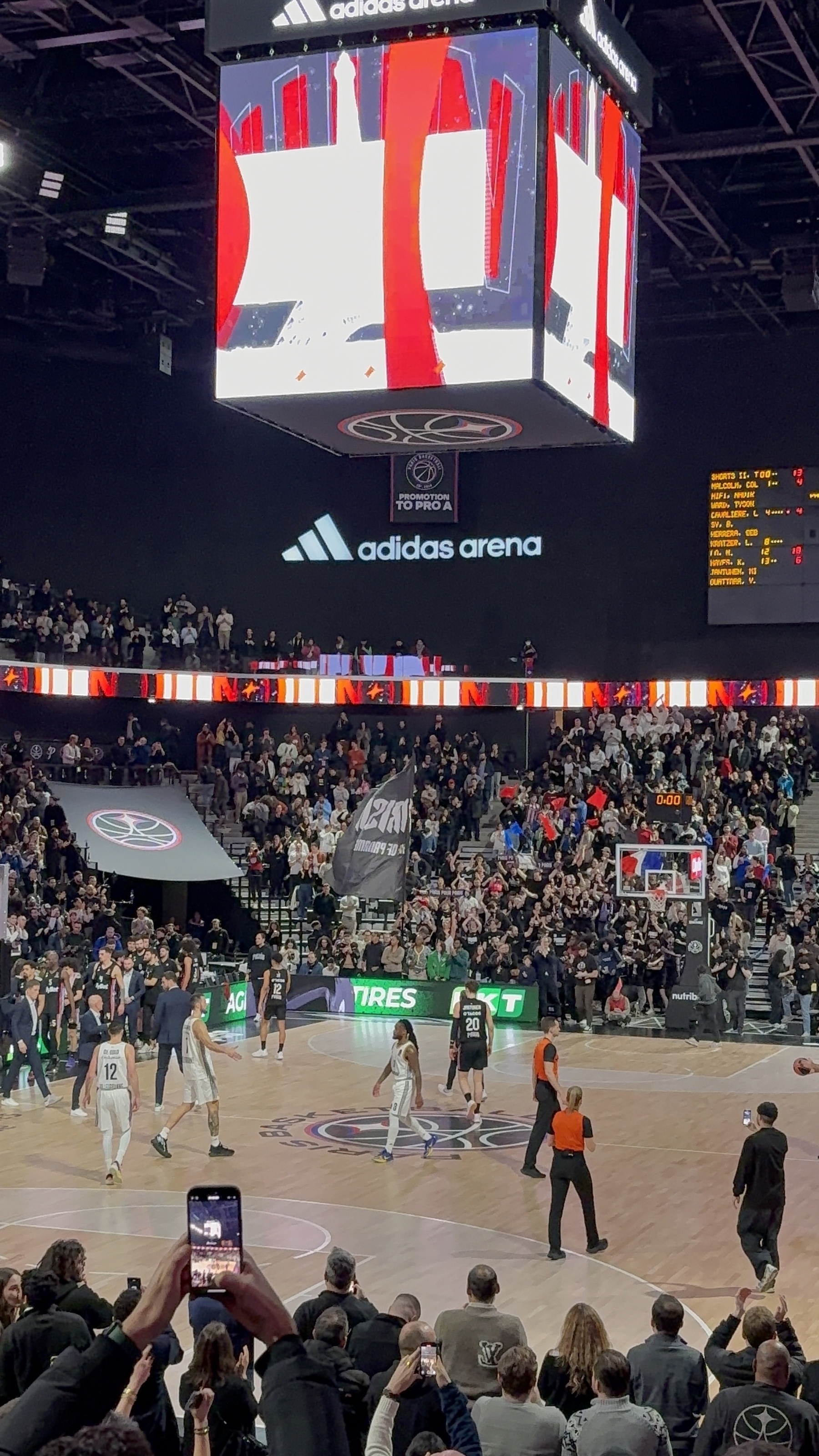 A basketball game is taking place in an arena with a large crowd and electronic displays, featuring players on the court and spectators capturing the moment on their phones.