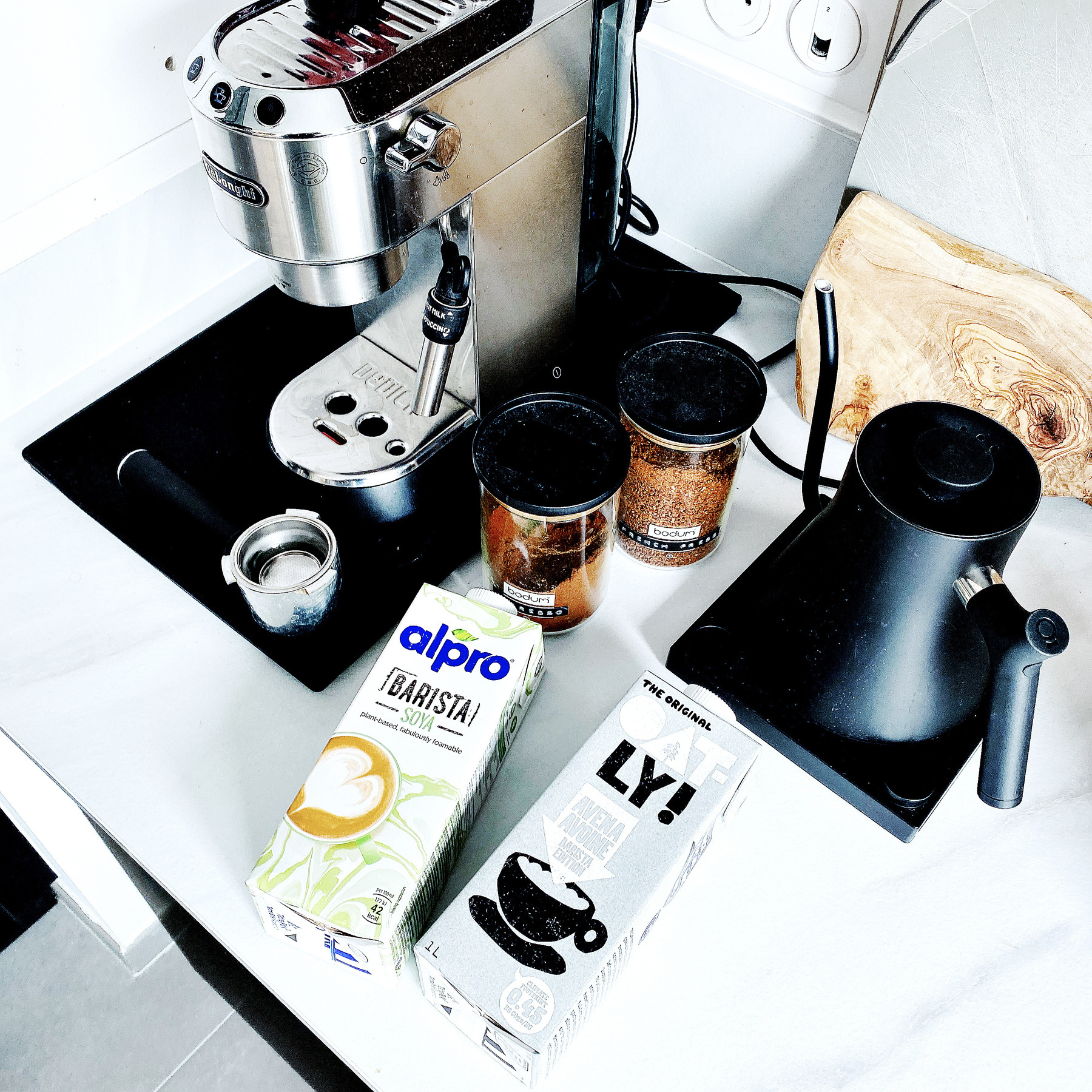 Auto-generated description: A coffee setup featuring a grinder, milk frothers, two types of plant-based milk, and a kettle is displayed on a countertop.