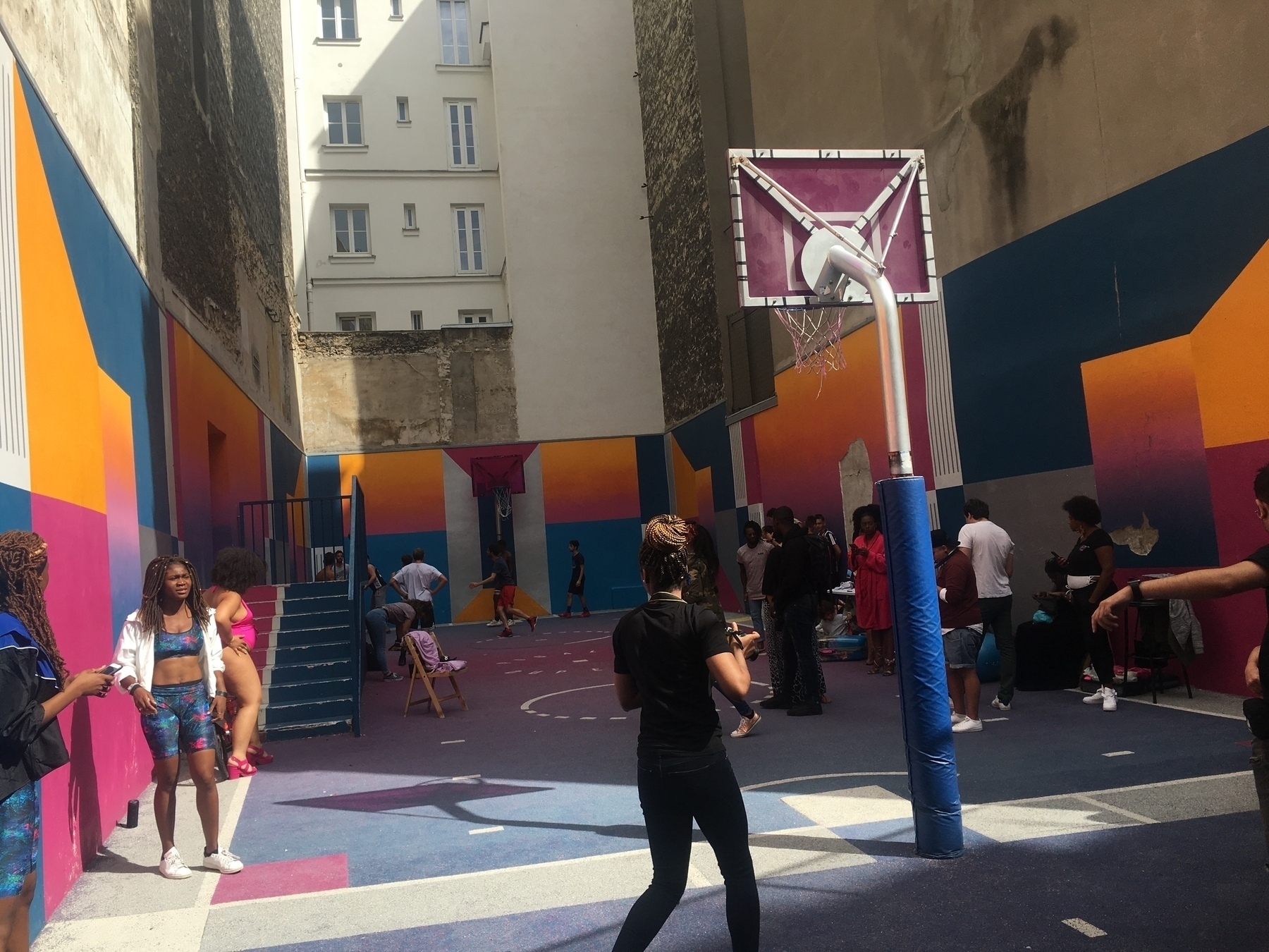 A group of people are gathered in a colorful outdoor basketball court surrounded by tall buildings.