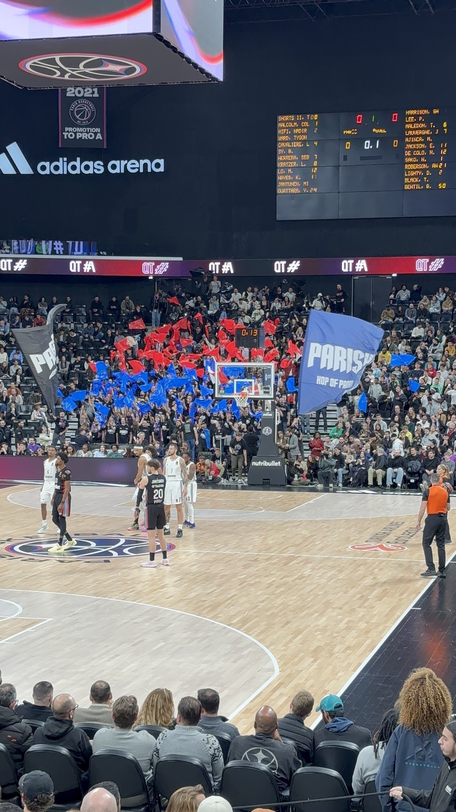 Basketball players are on the court during a game at a packed arena with fans waving flags and banners.