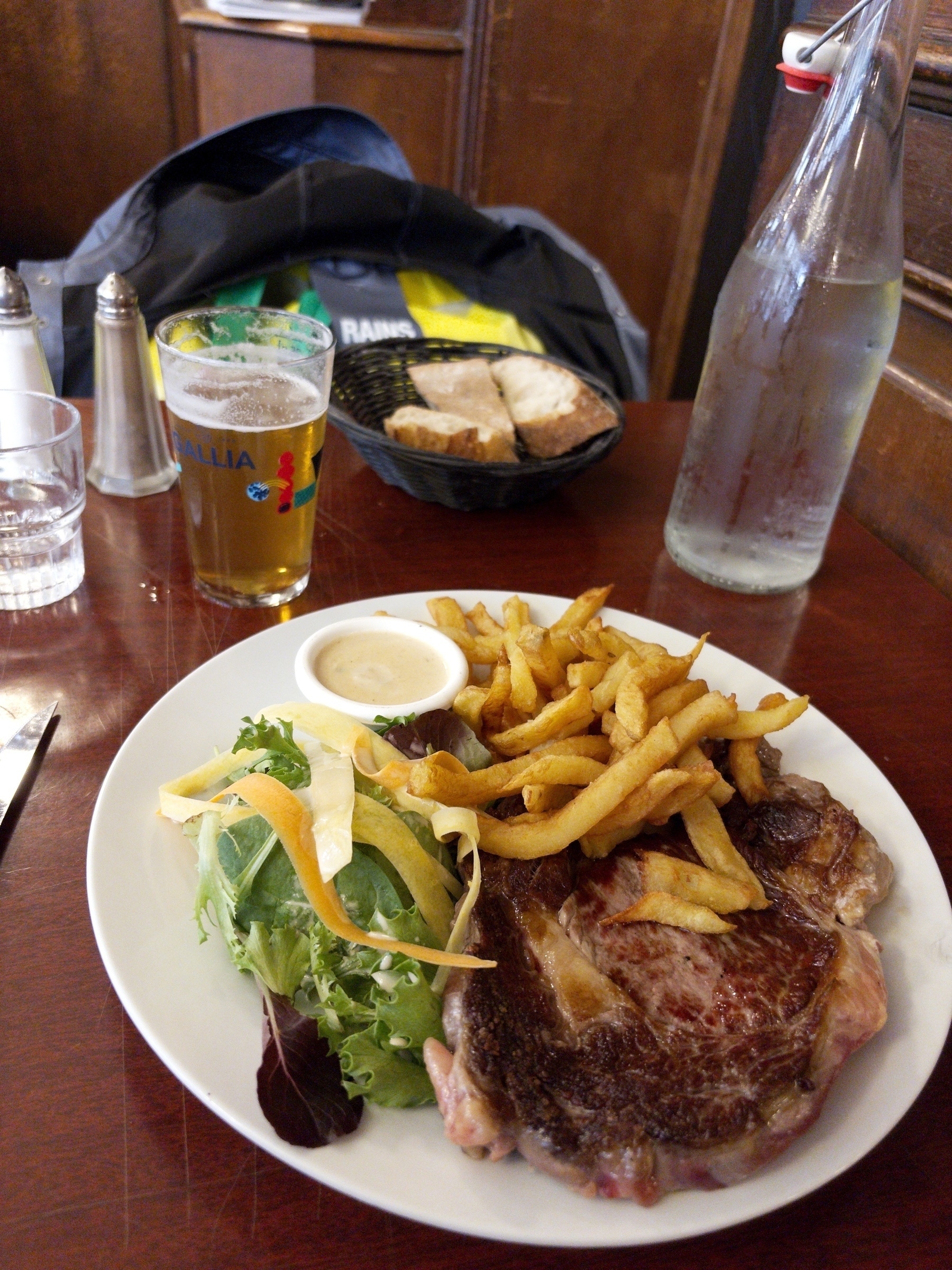 Auto-generated description: A plate with steak, fries, and salad is on a table alongside a glass of beer, water bottle, and a basket of bread.