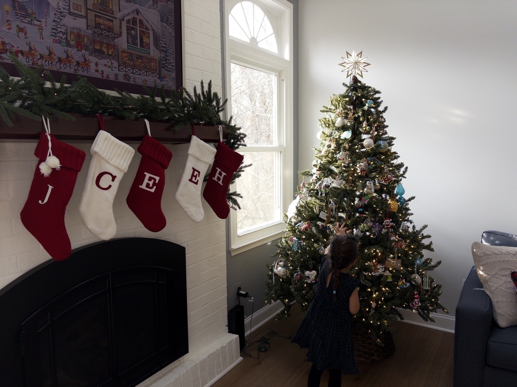 A decorated Christmas tree stands beside a fireplace with stockings and a child admiring the tree.