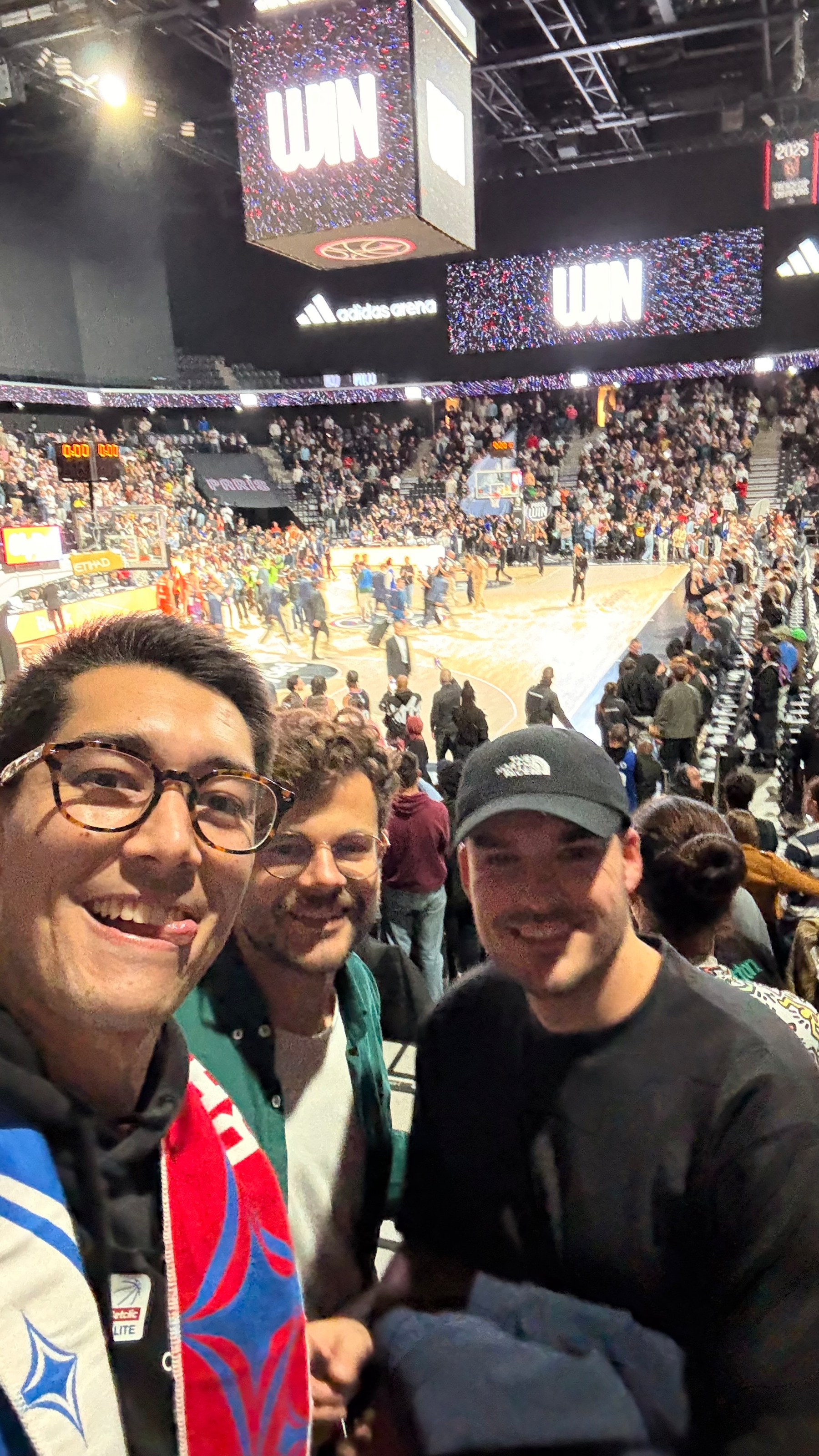 Three people are smiling in the foreground of a crowded basketball arena.