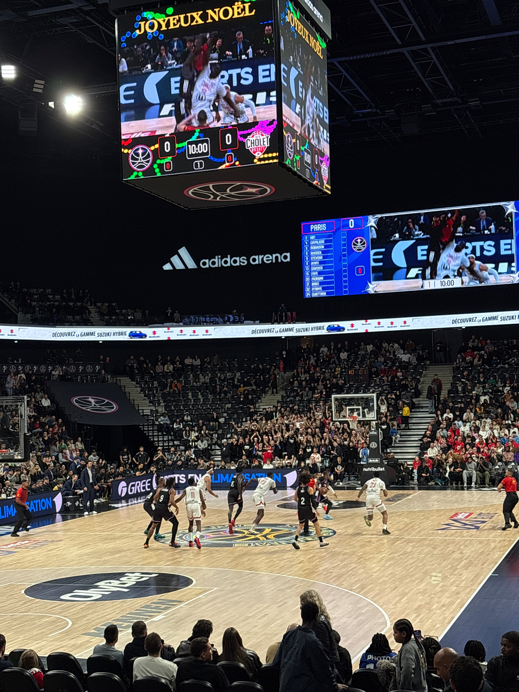 A basketball game is taking place in a large indoor arena with spectators, featuring displays showing holiday greetings and team scores.