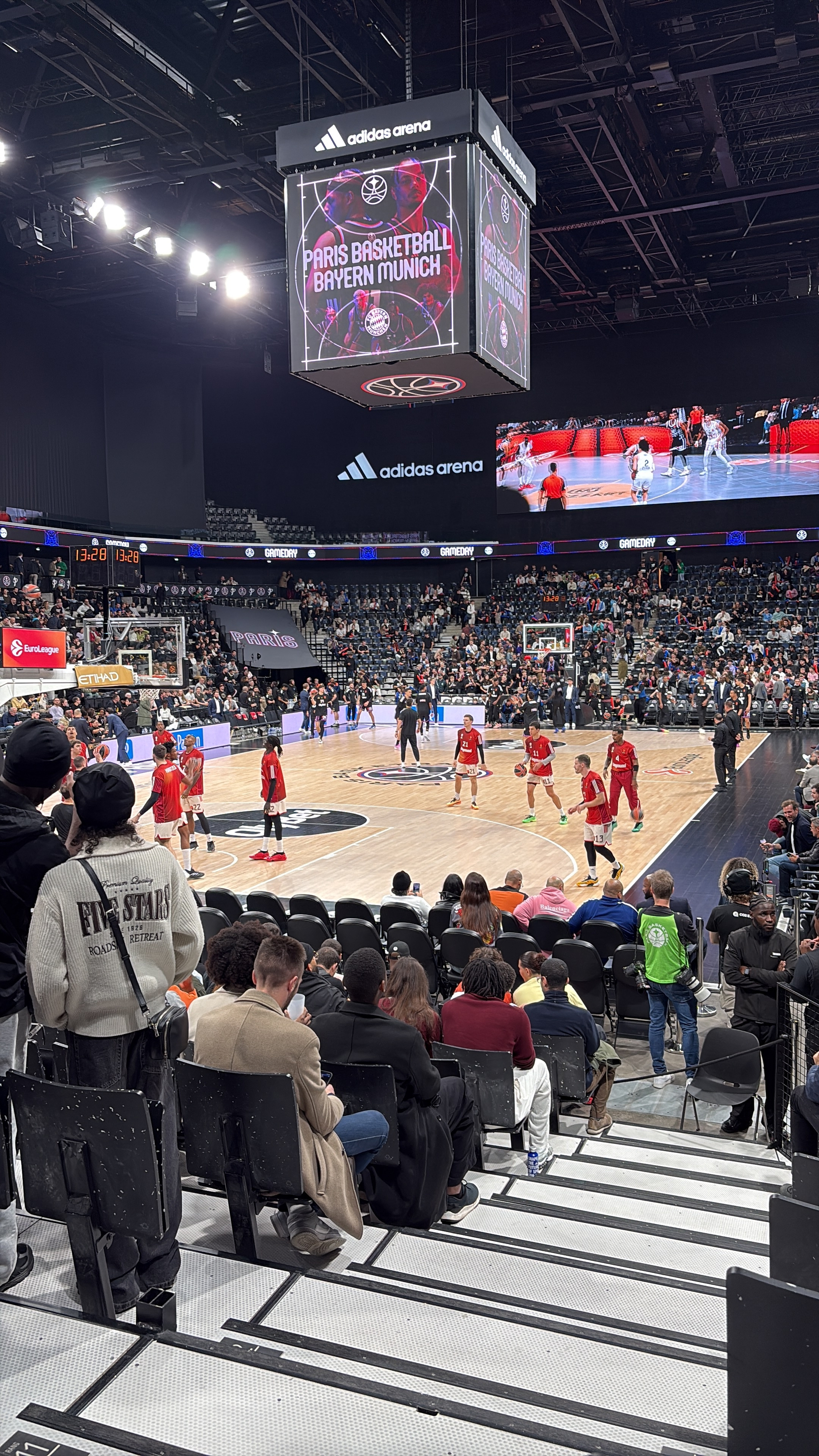 A basketball game is taking place in a large indoor arena with players in red and white uniforms on the court, watched by a sizable crowd.