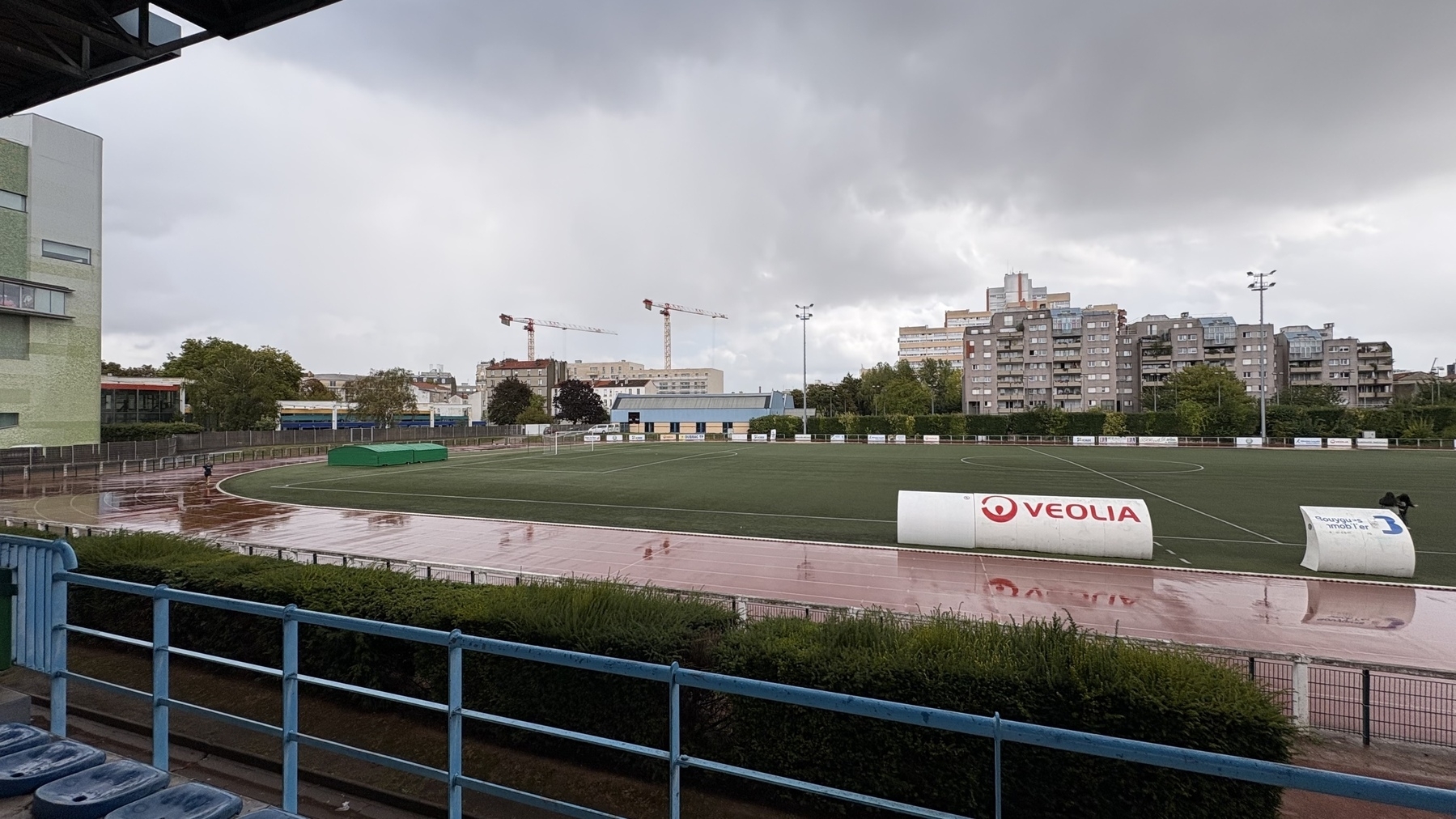 A sports field with a running track is set against a backdrop of cranes and apartment buildings, under a cloudy sky.