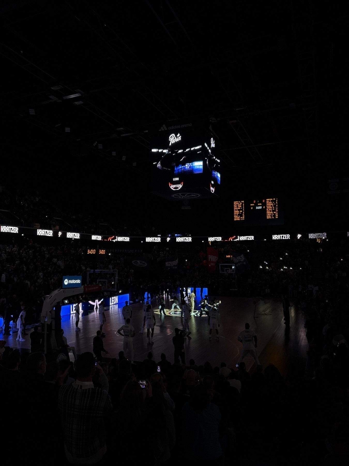 A basketball game is taking place in a dimly lit arena with players and an audience visible.