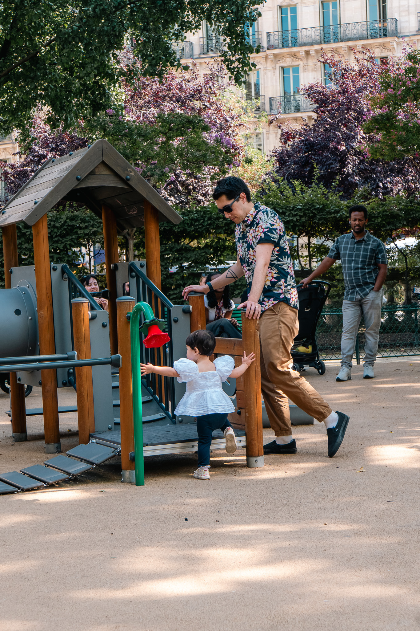 A man is helping a young child at a playground with a play structure nearby, while other people and trees are in the background.