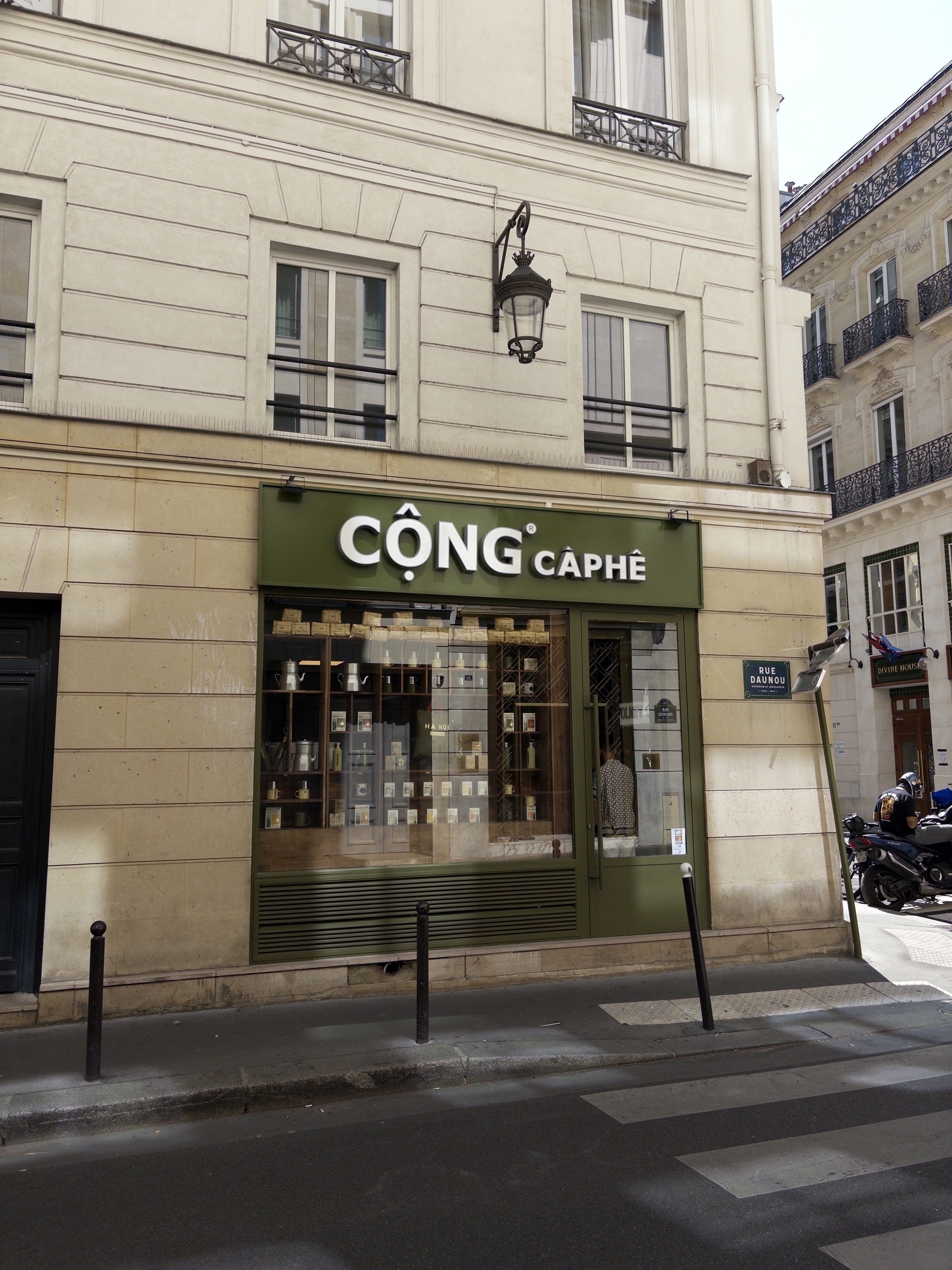 A Cộng Cà Phê coffee shop is situated on the corner of a street with beige buildings in the background.
