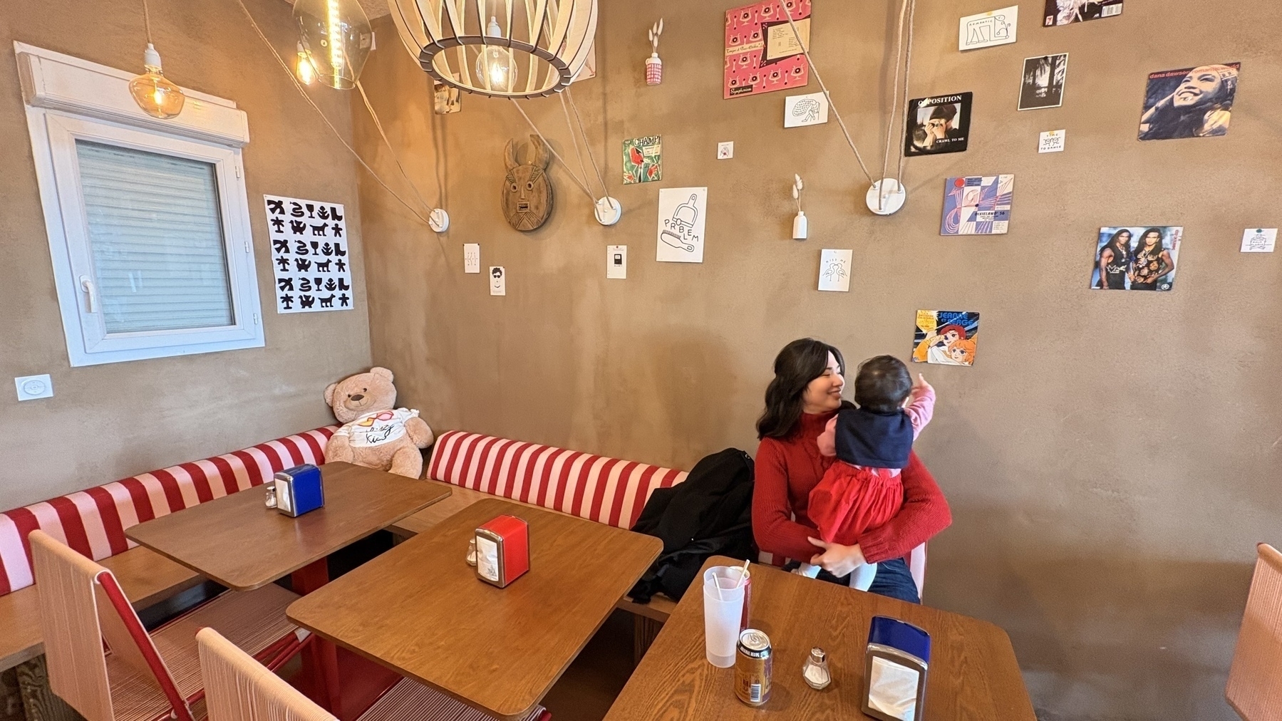 A woman is sitting with a baby in a cafe adorned with various wall decorations and a large teddy bear on the bench.