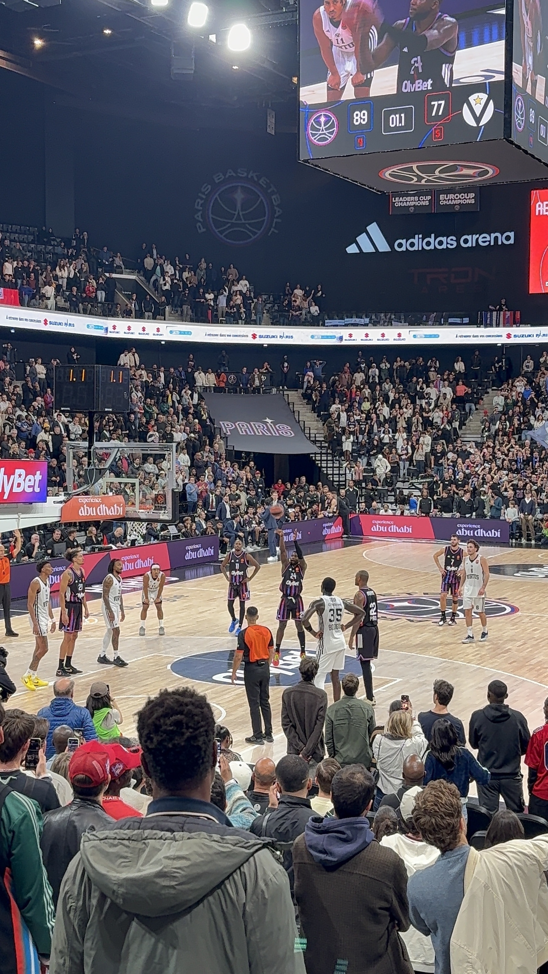 A basketball game is taking place in a packed arena with players on the court and a scoreboard displaying the score as 89-77.
