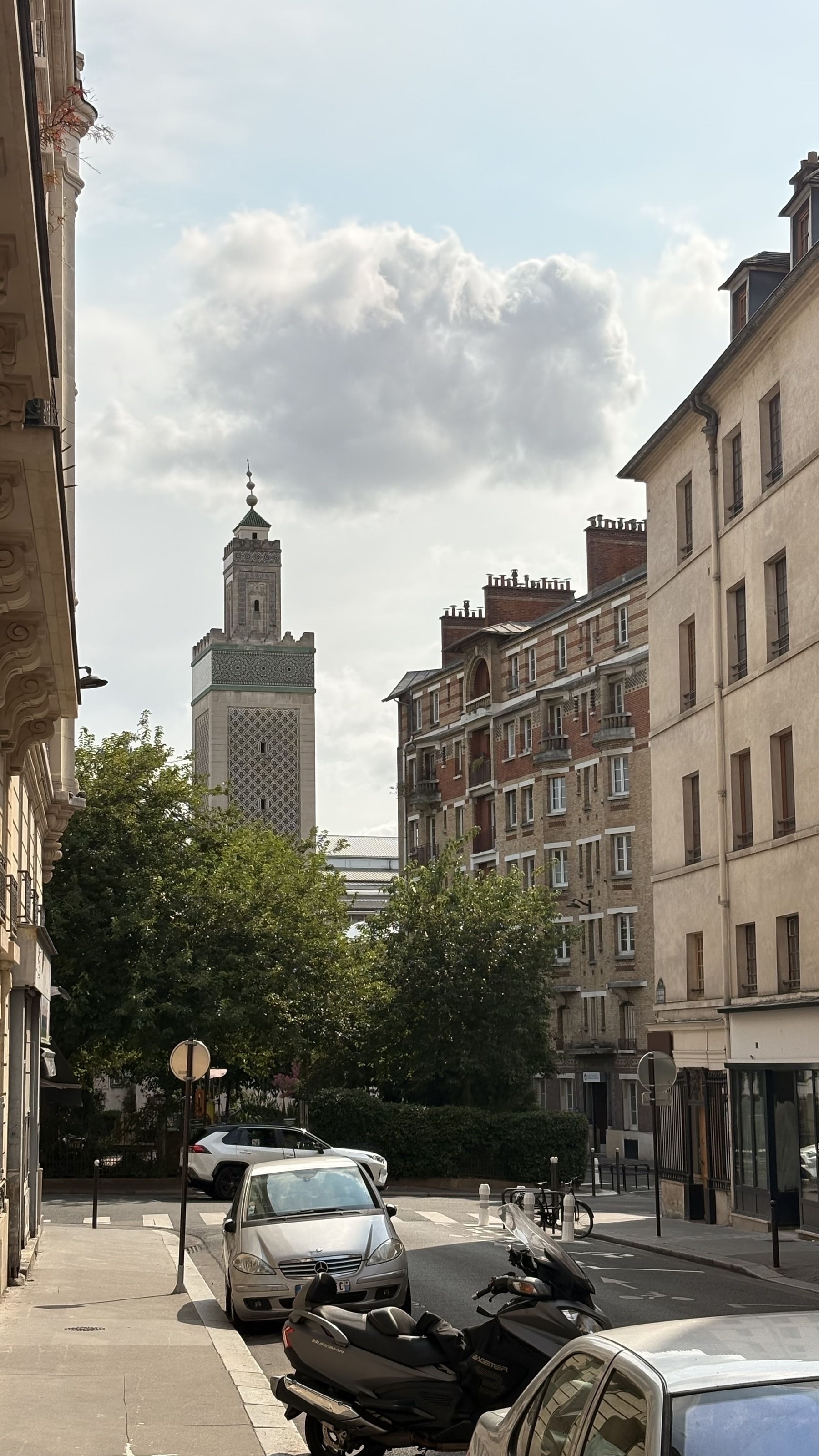 A quiet urban street lined with parked cars and scooters offers a view of a tall, ornate building in the background under a partly cloudy sky.