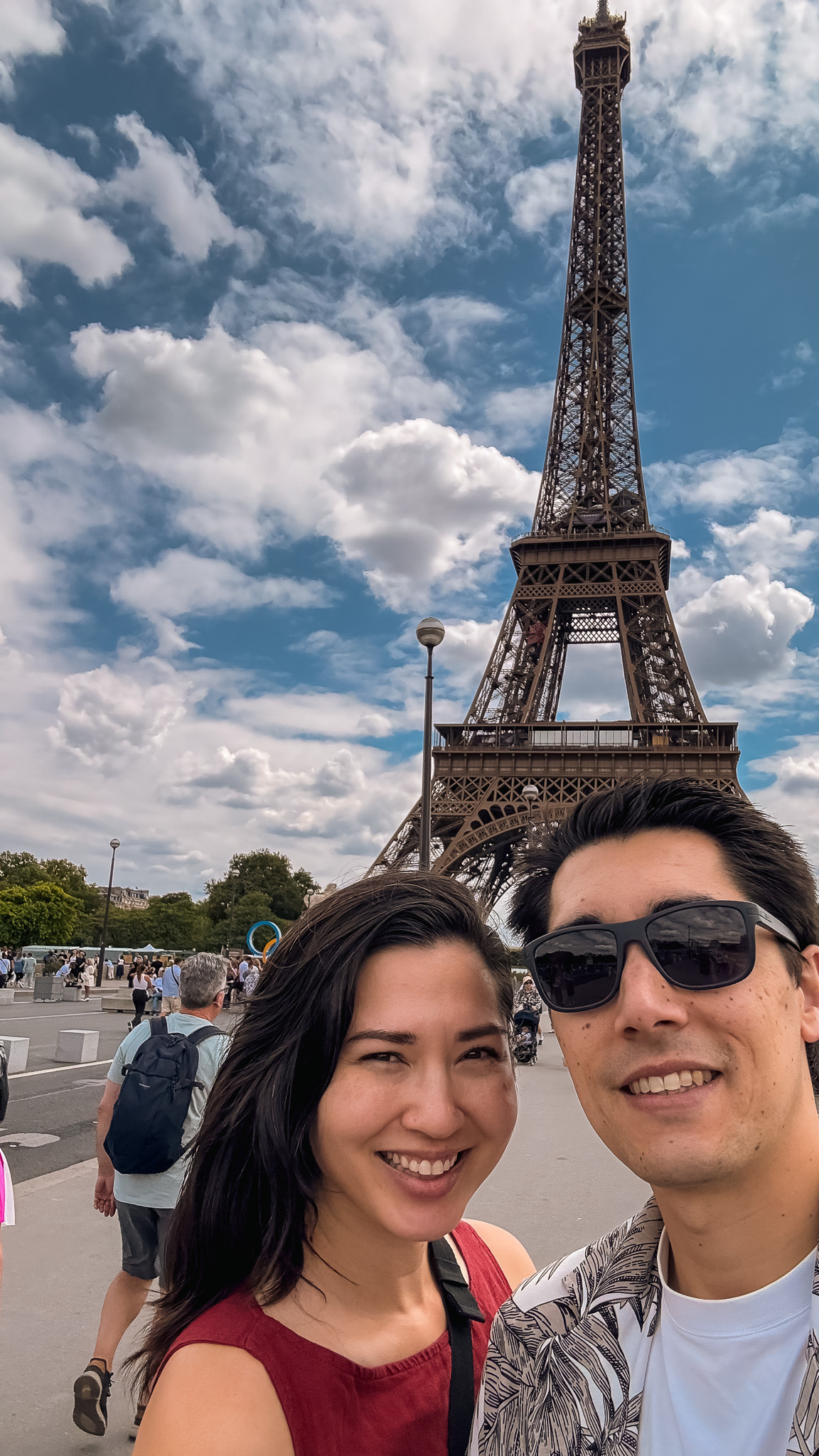 Two people are taking a selfie in front of the Eiffel Tower on a sunny day.