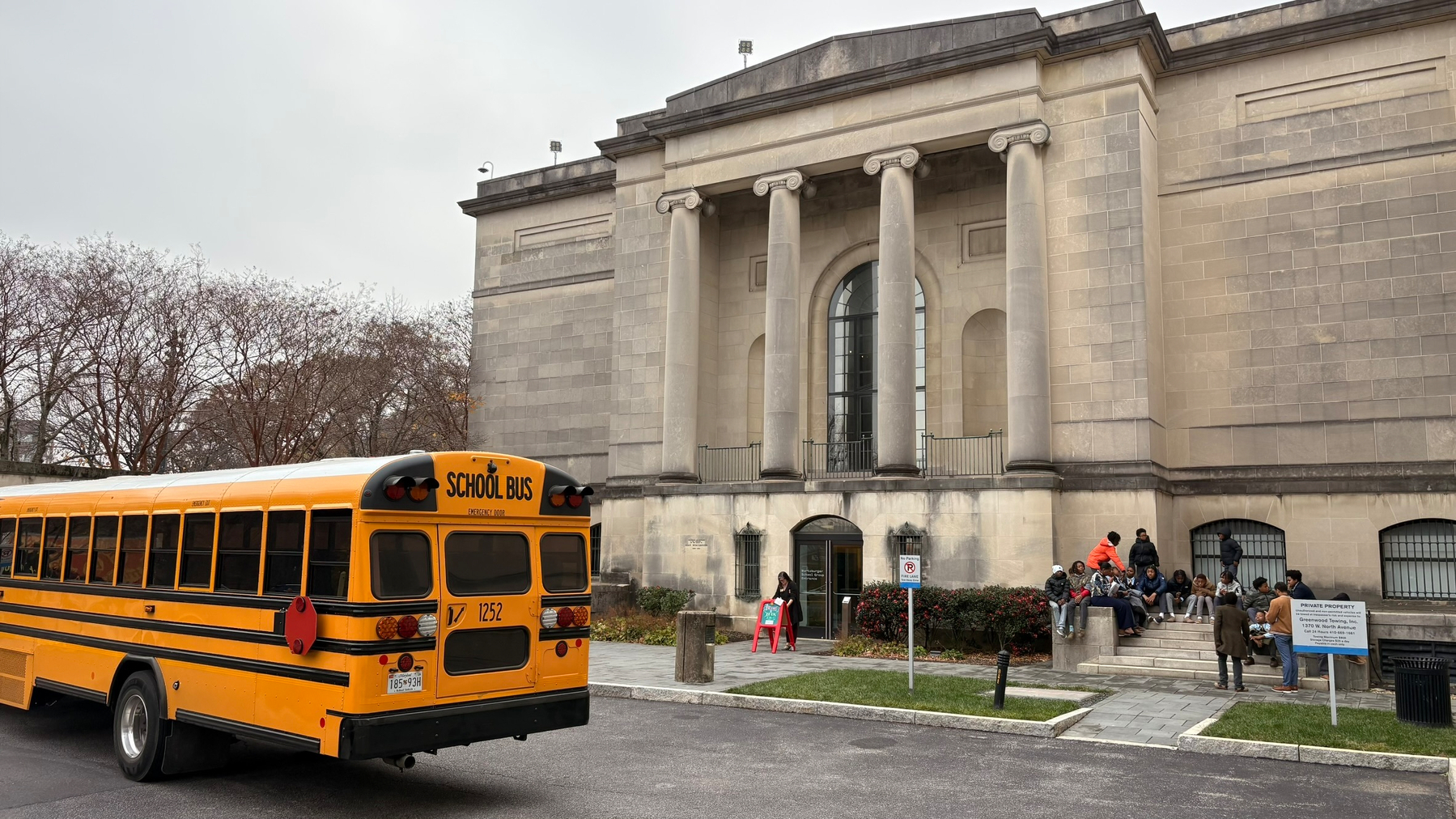 A yellow school bus is parked in front of a large, historic-looking building with columns, while a group of people gather near the entrance.