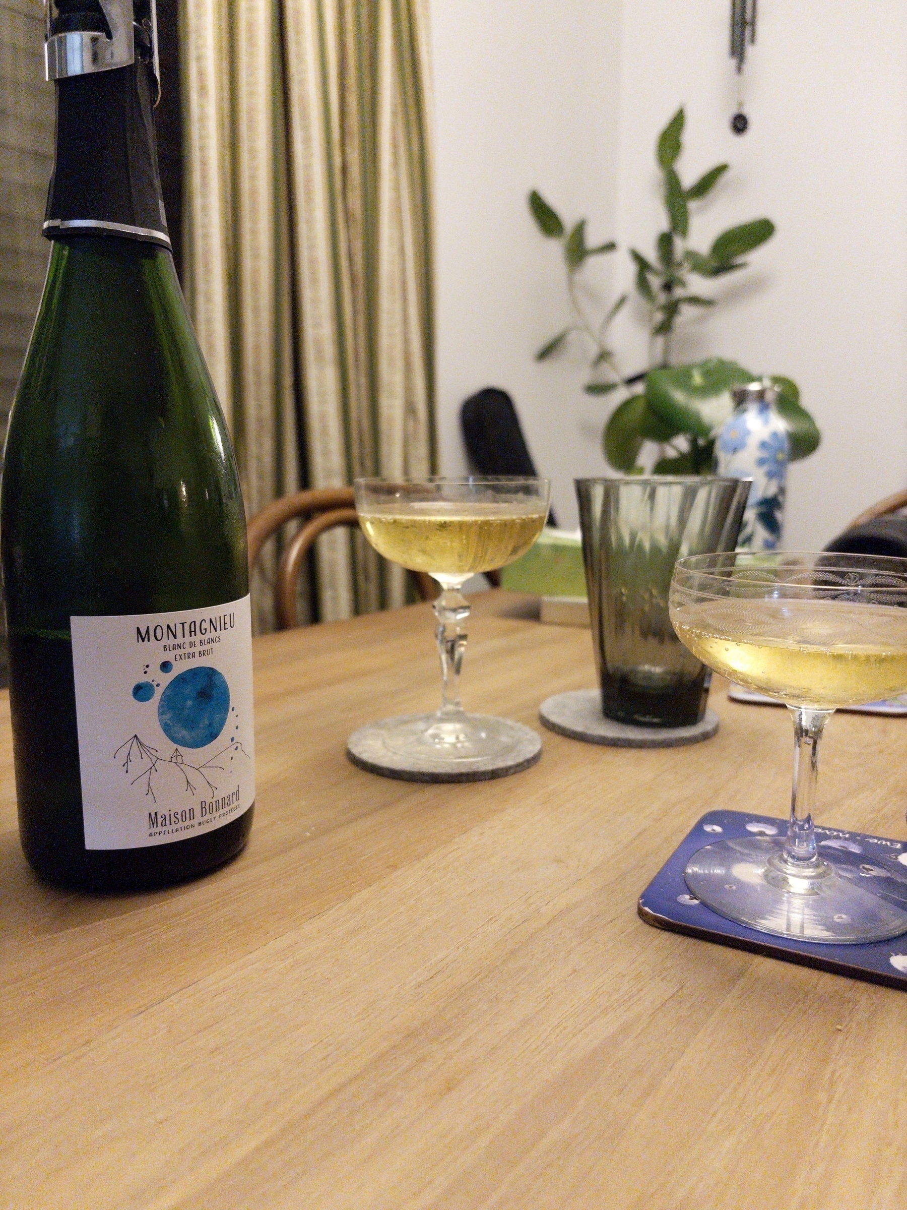 A bottle of sparkling wine labeled "Maison Briard" is on a wooden table with two filled coupe glasses and a green glass, set against a backdrop of curtains and a plant.