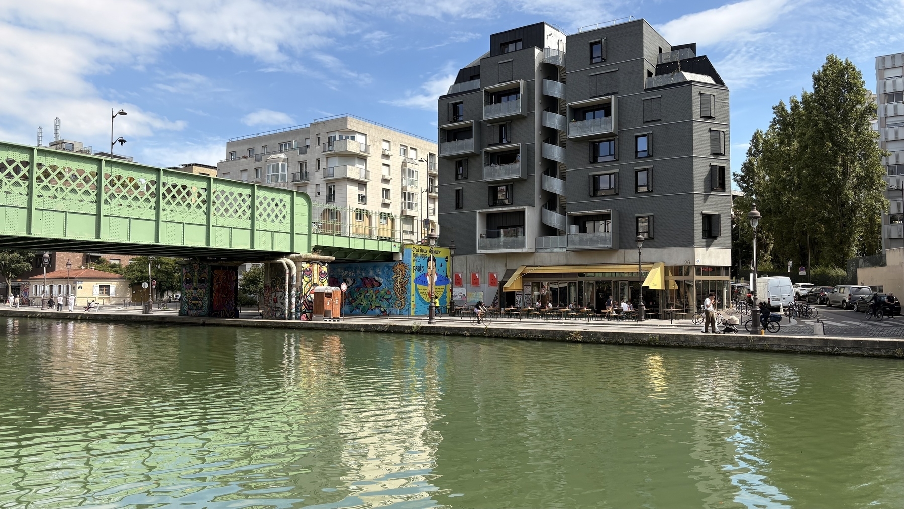 A green iron bridge stretches over a canal beside modern buildings, with a mural and a café visible along the waterfront.