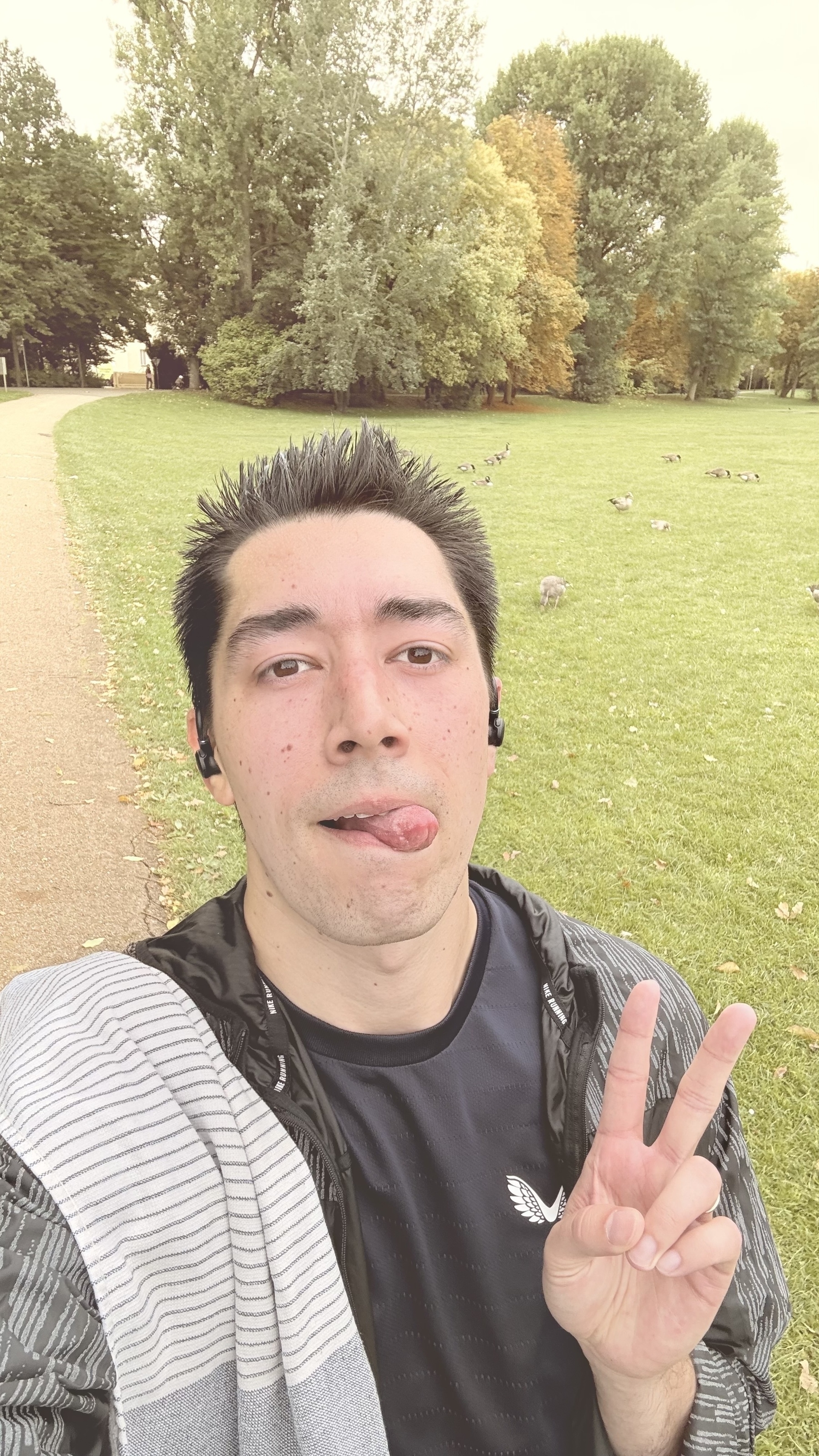 A person holding up a peace sign poses for a selfie in a park, wearing a striped towel and earbuds.