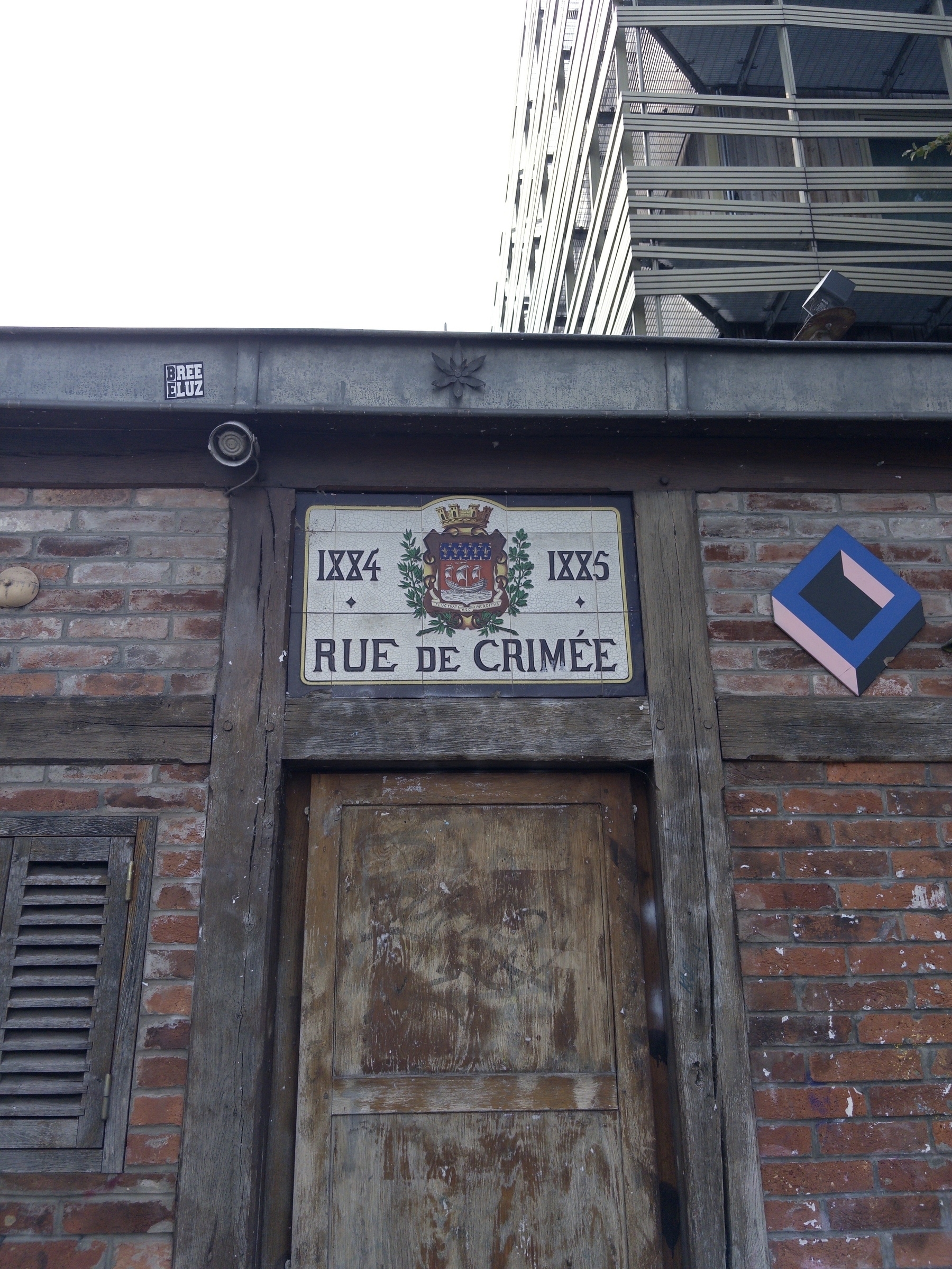 A weathered brick wall features a wooden door and a street sign labeled ‘Rue de Crimée’ with a building in the background.