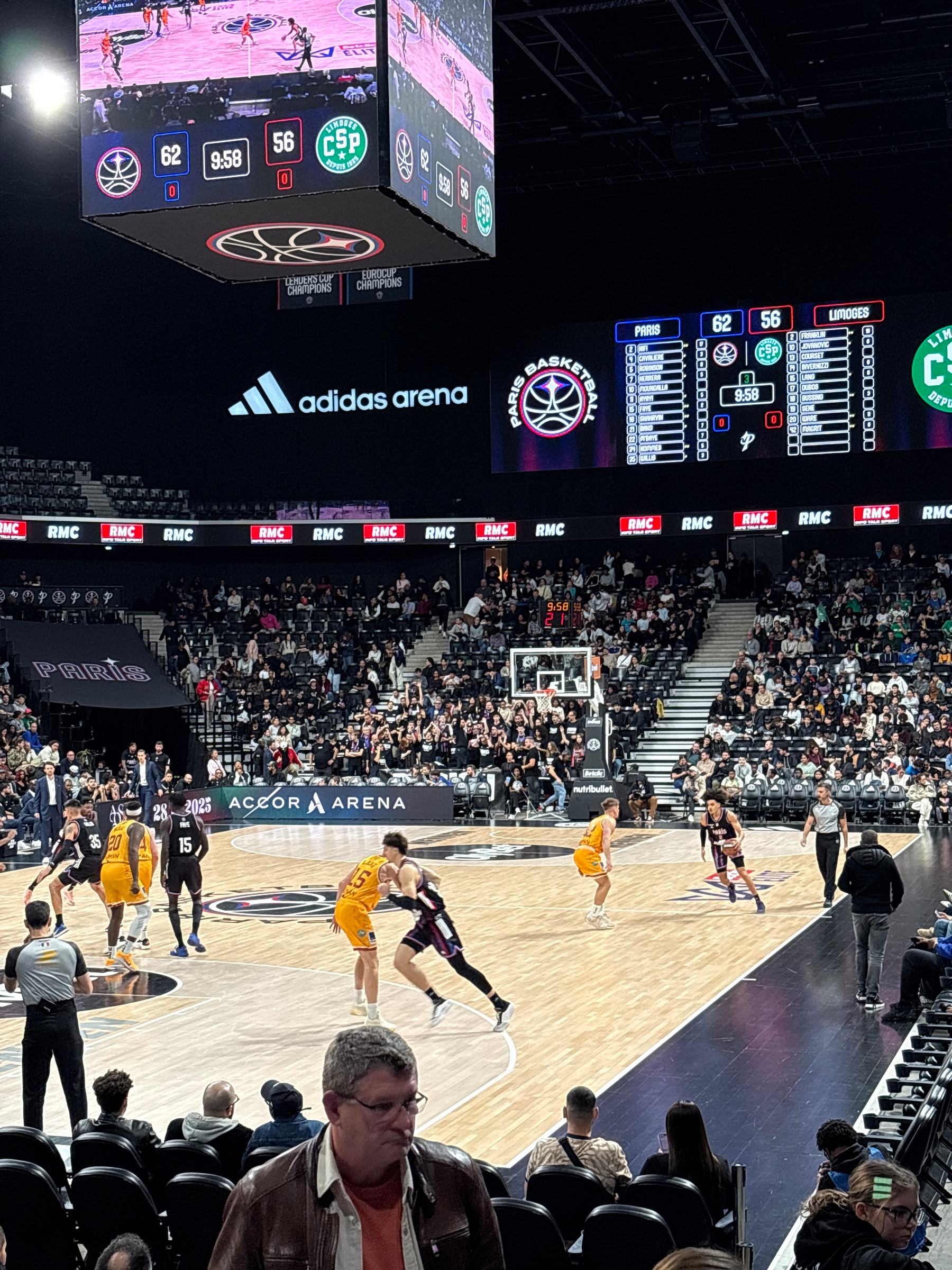 A basketball game is taking place in a large indoor arena with a scoreboard displaying the scores and crowd in attendance.