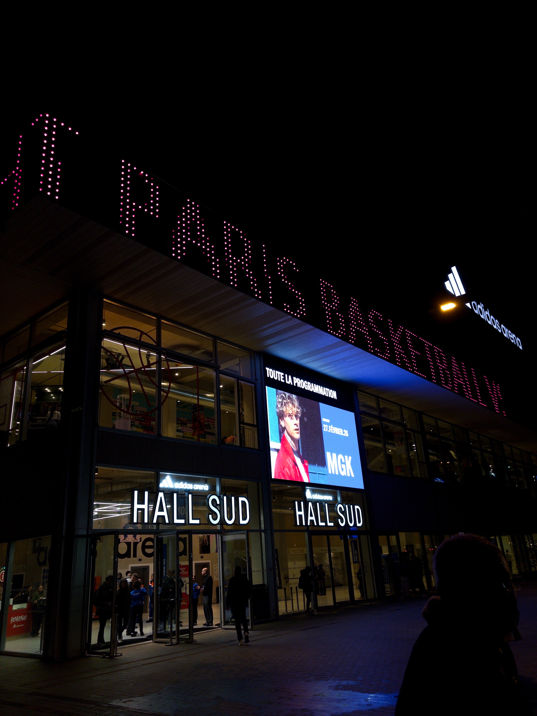 A nighttime scene showcases an illuminated basketball stadium entrance with people entering and a large digital screen displaying event information.