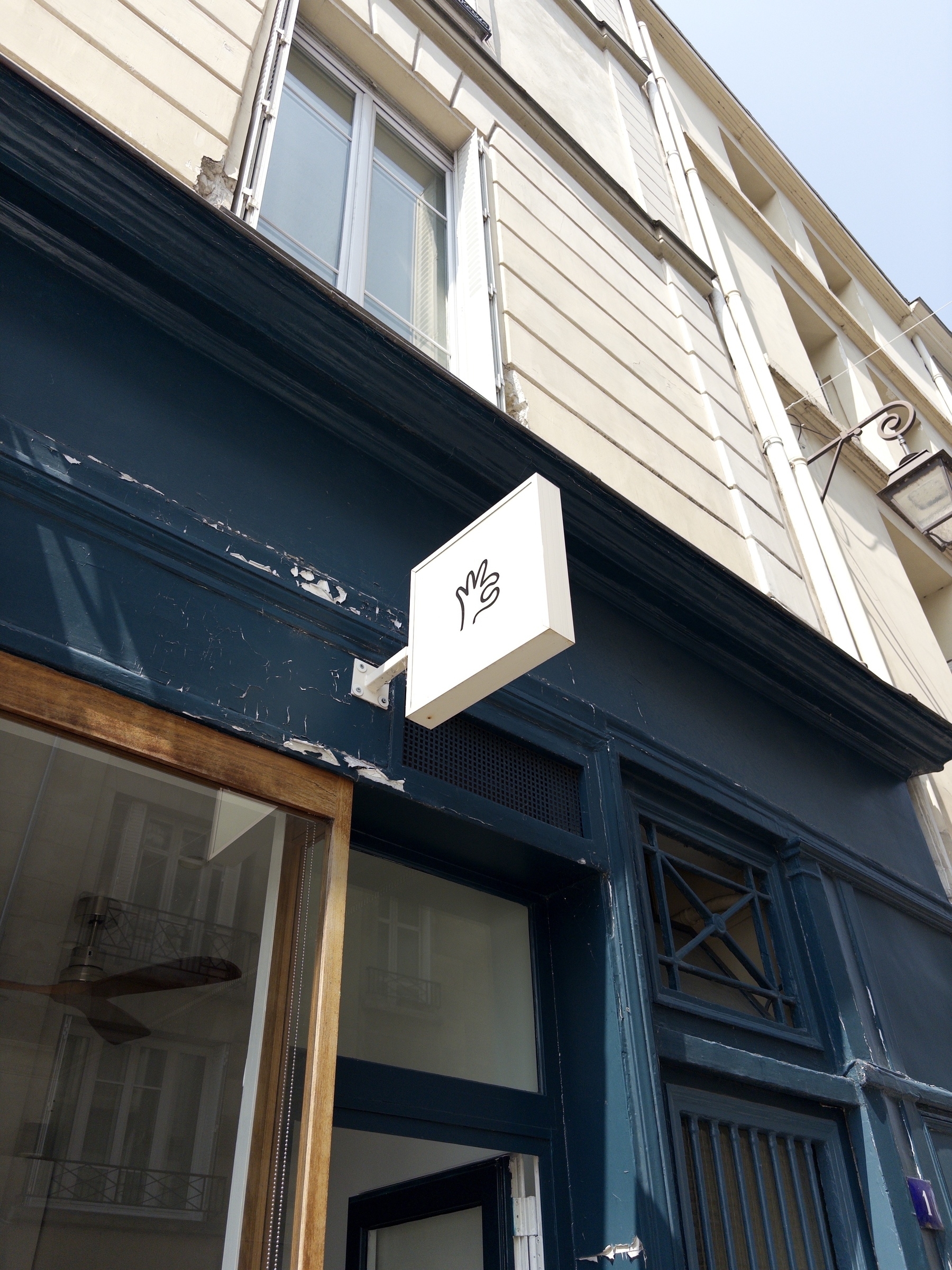 A simple storefront sign featuring a drawing of a hand is mounted on the corner of a building with blue and beige accents.