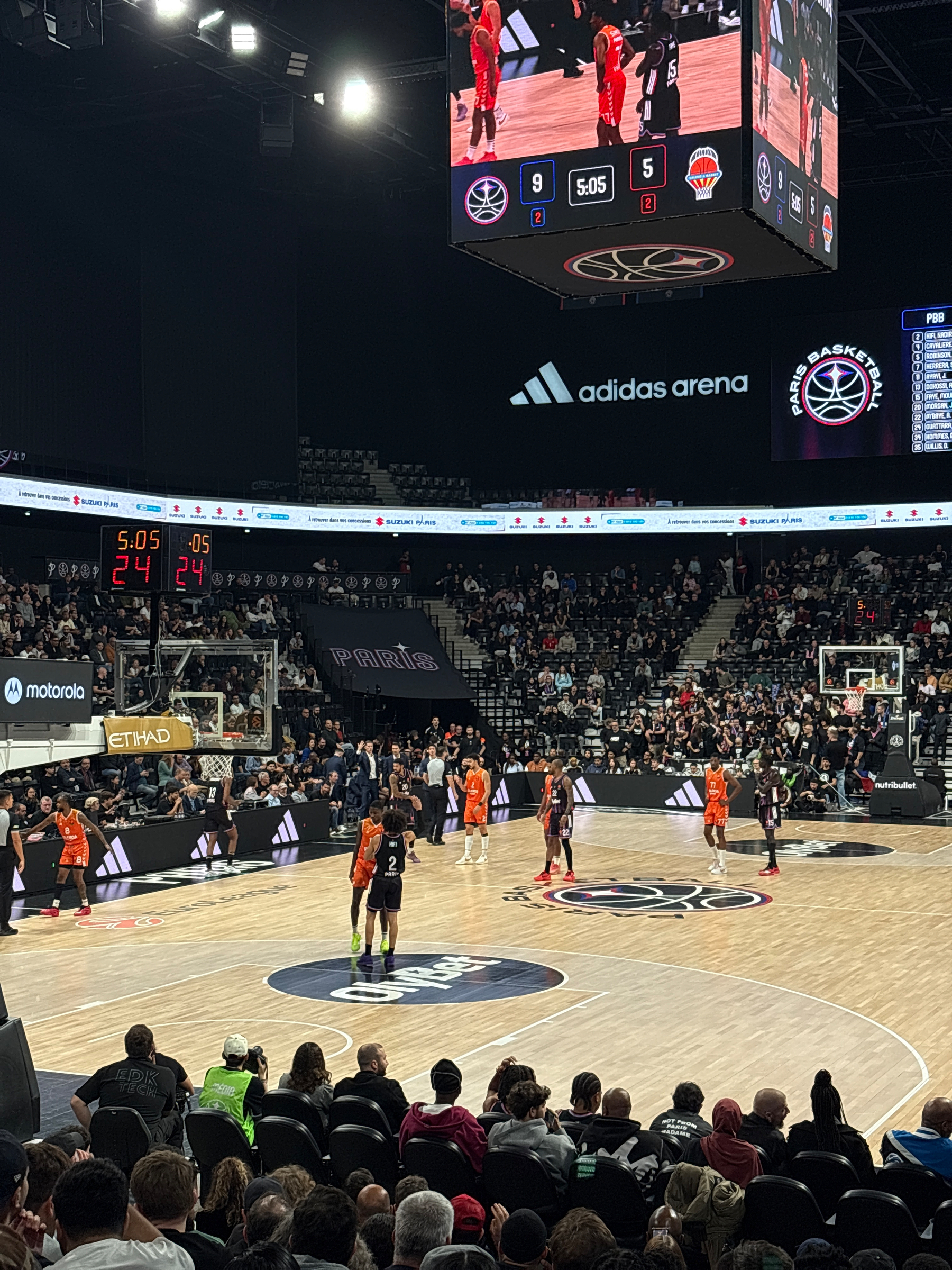 A basketball game is taking place in an indoor arena with spectators watching from the stands.
