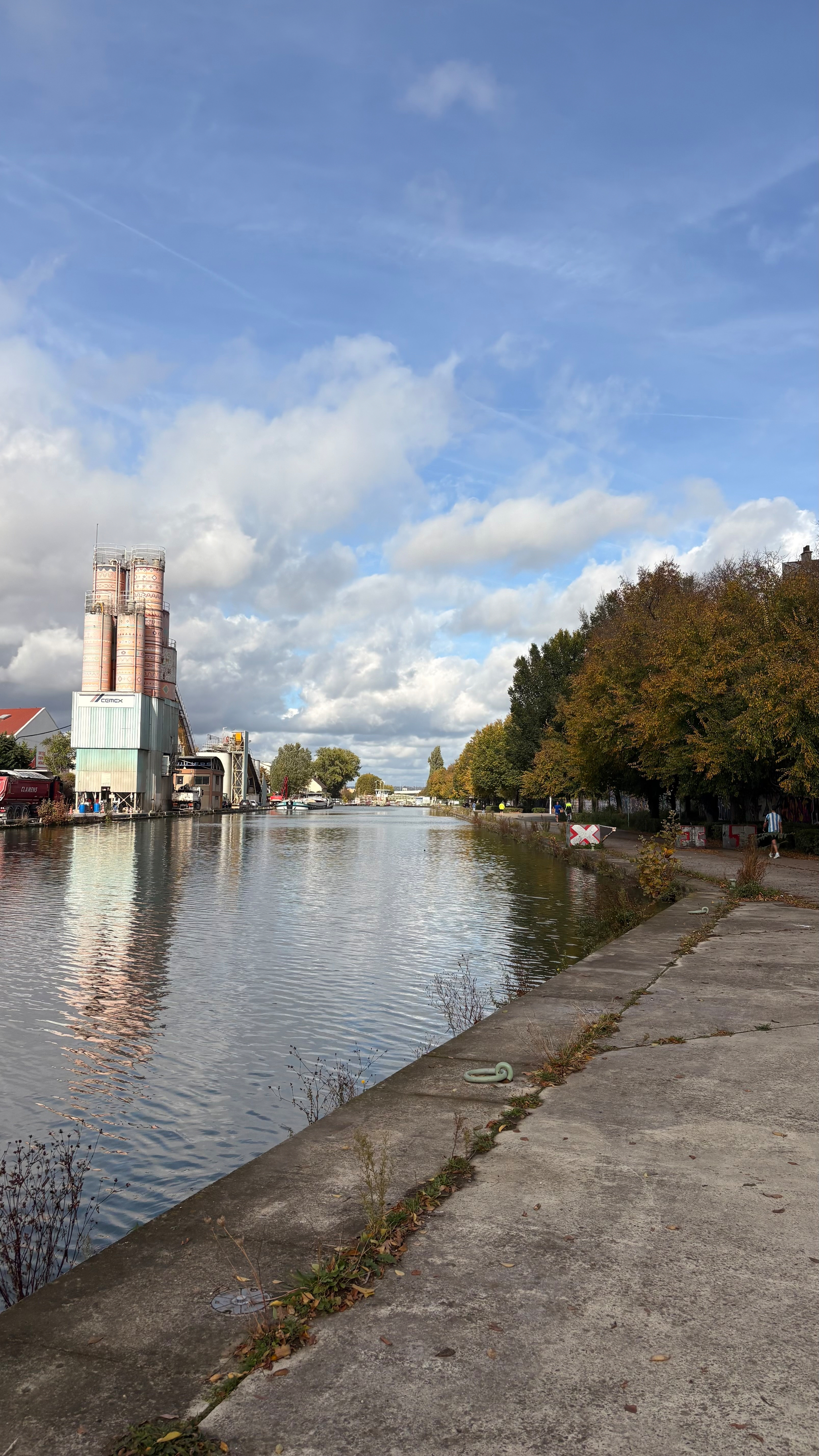 A serene riverside walkway is flanked by a large industrial building on the left and trees with autumn foliage on the right, under a partly cloudy sky.