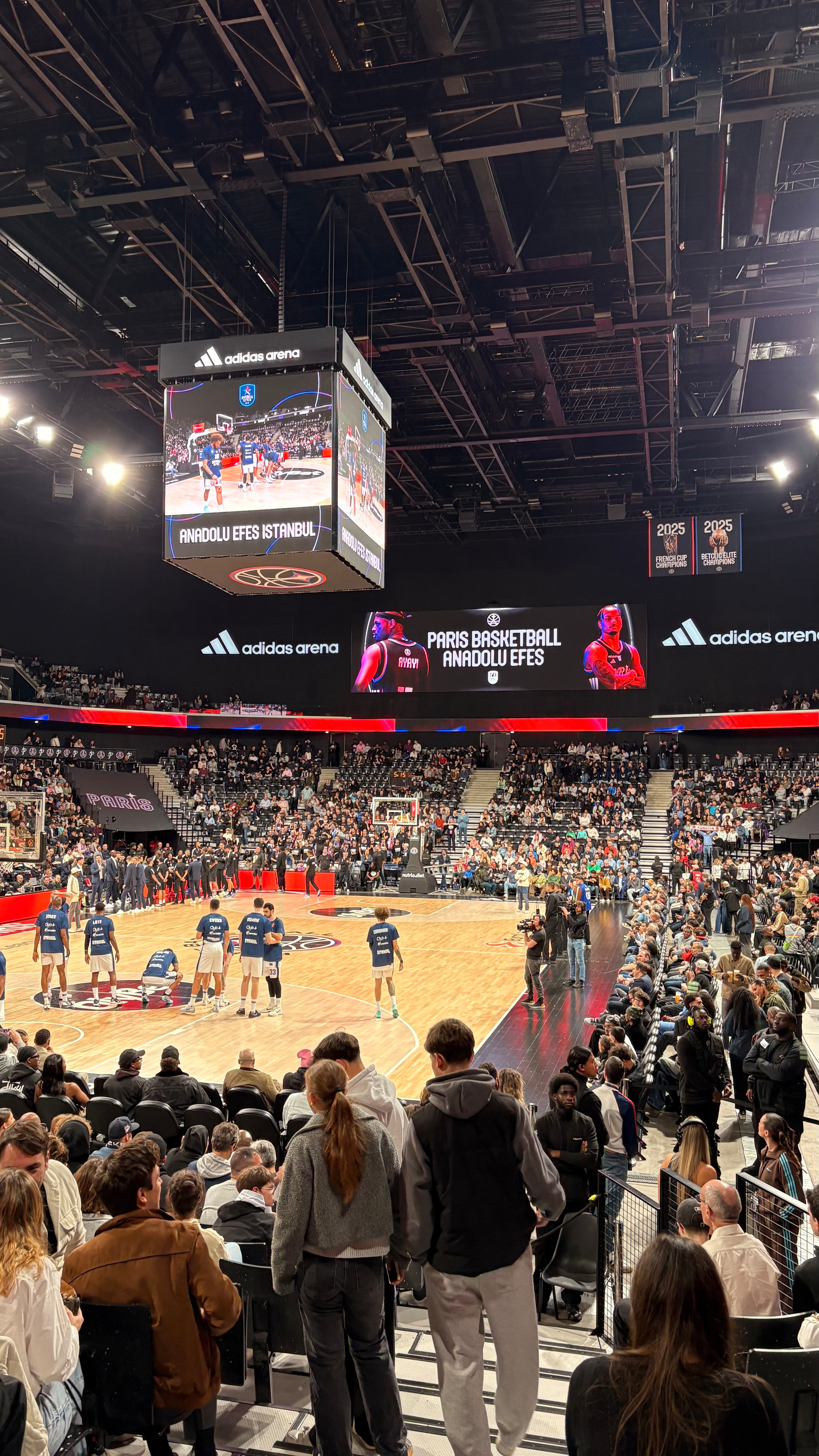 A basketball game is taking place in an indoor arena with spectators filling the seats and digital screens displaying game information.
