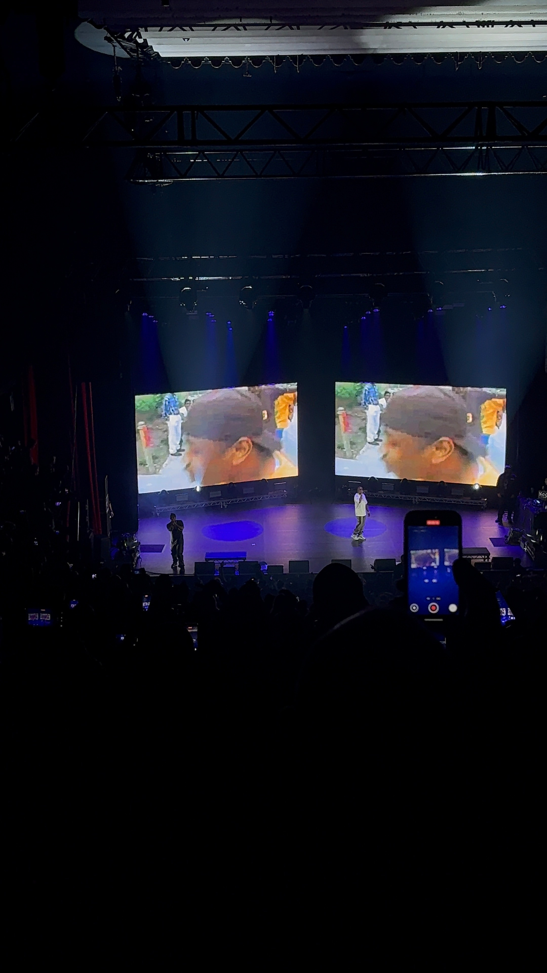 A concert stage is illuminated with blue lights, featuring two large screens displaying a video, while silhouettes of the audience are visible in the foreground.