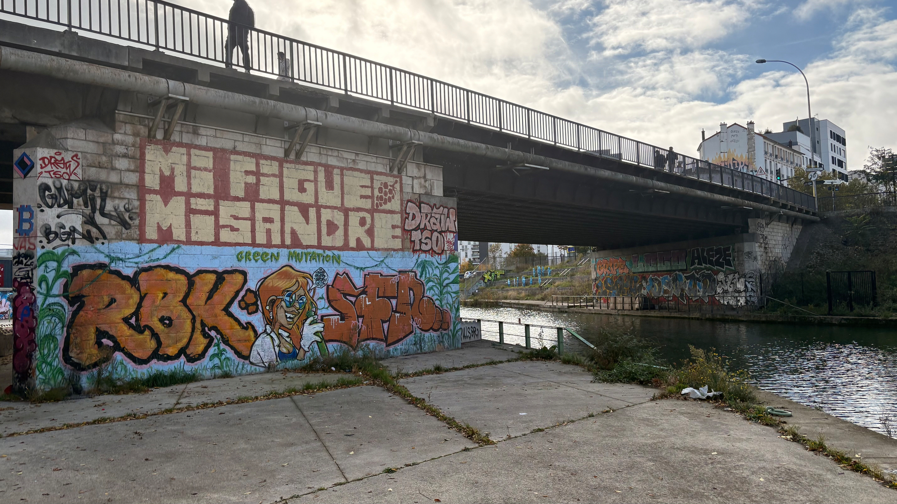 A graffiti-covered bridge spans over a canal, with various colorful tags and designs visible on the concrete surfaces.