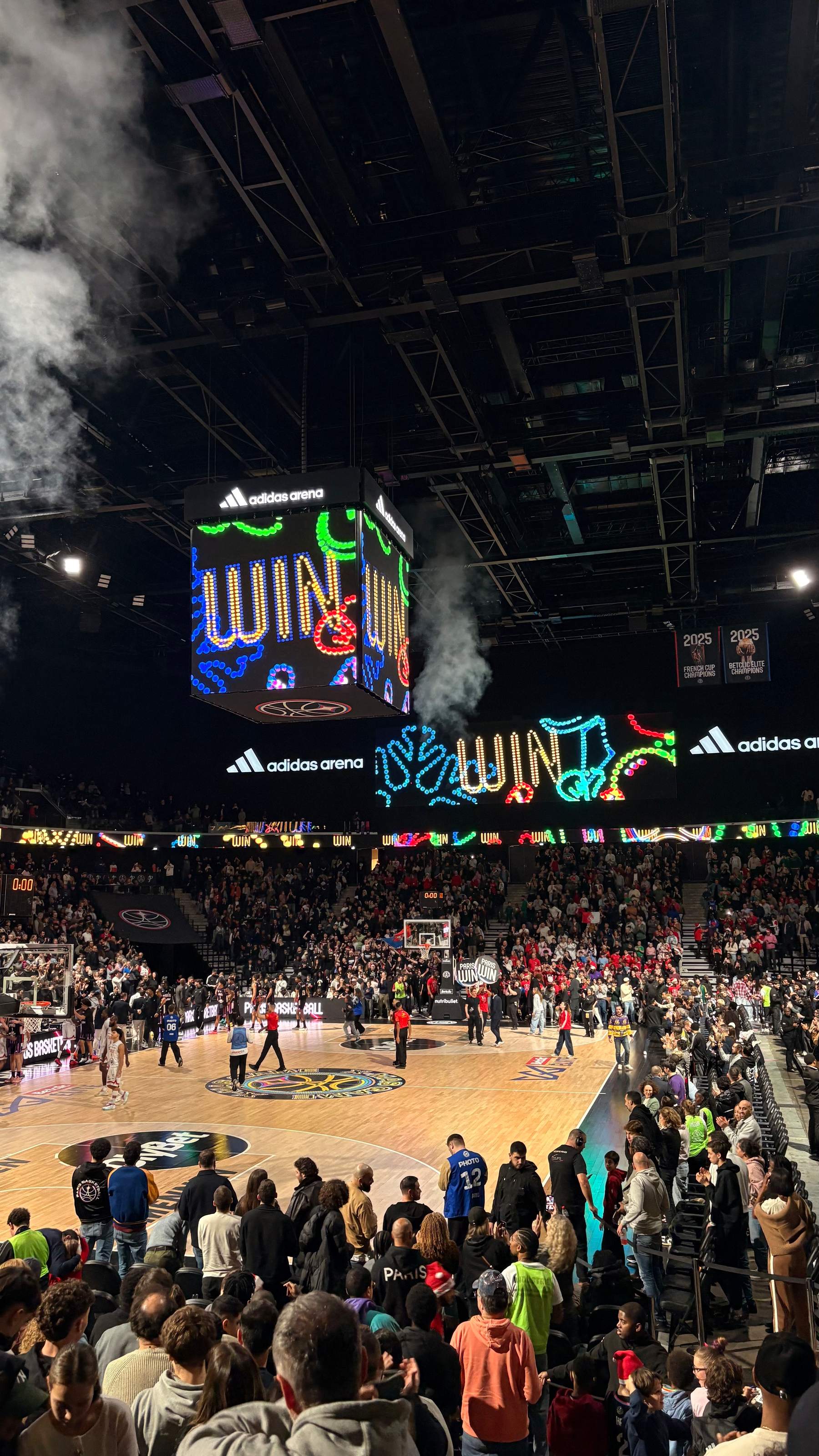 A basketball game is taking place in an indoor arena with a large screen displaying celebratory graphics and the word WIN.