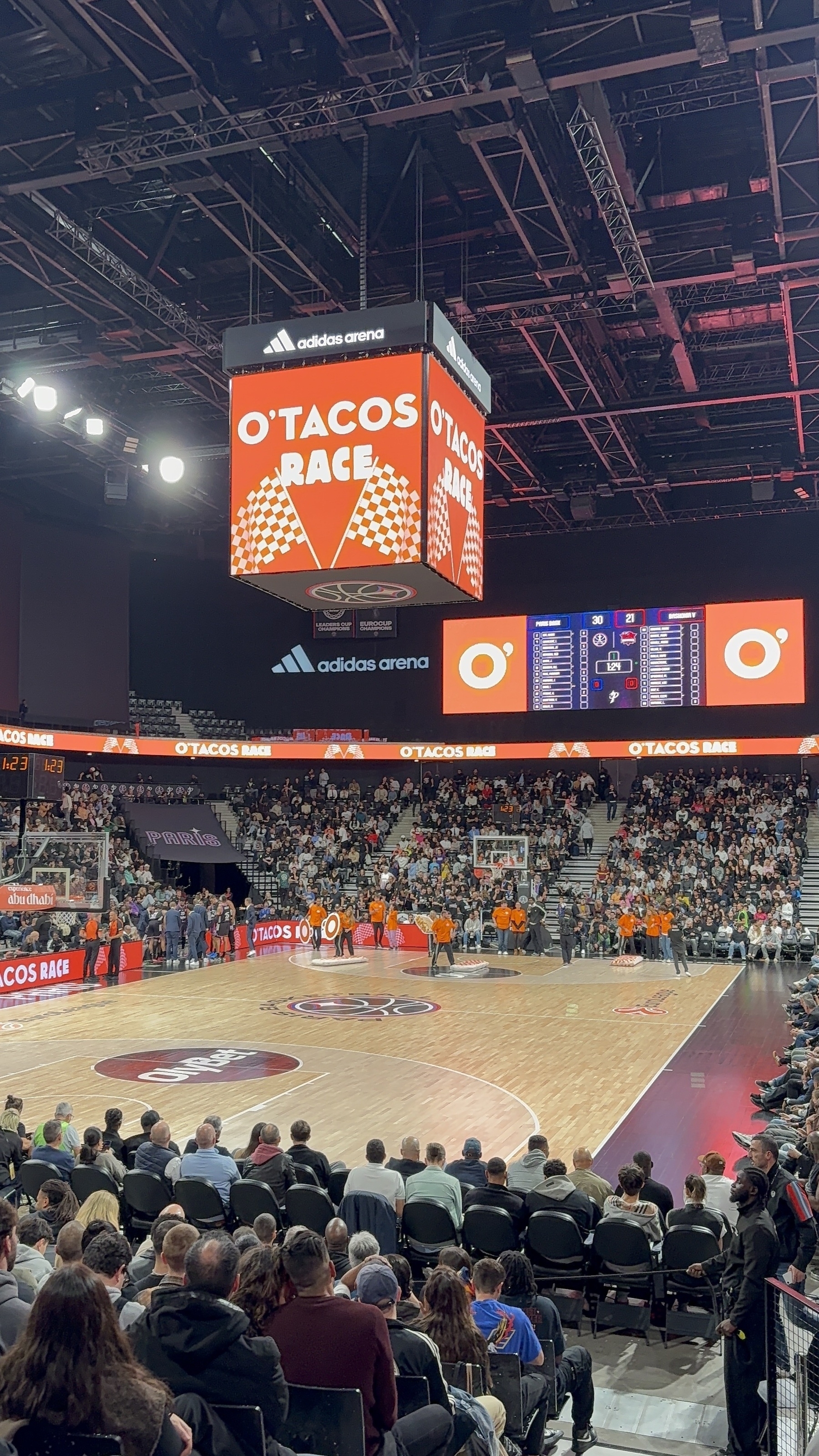 A basketball game is taking place in an indoor arena with a large crowd and advertisements on display.