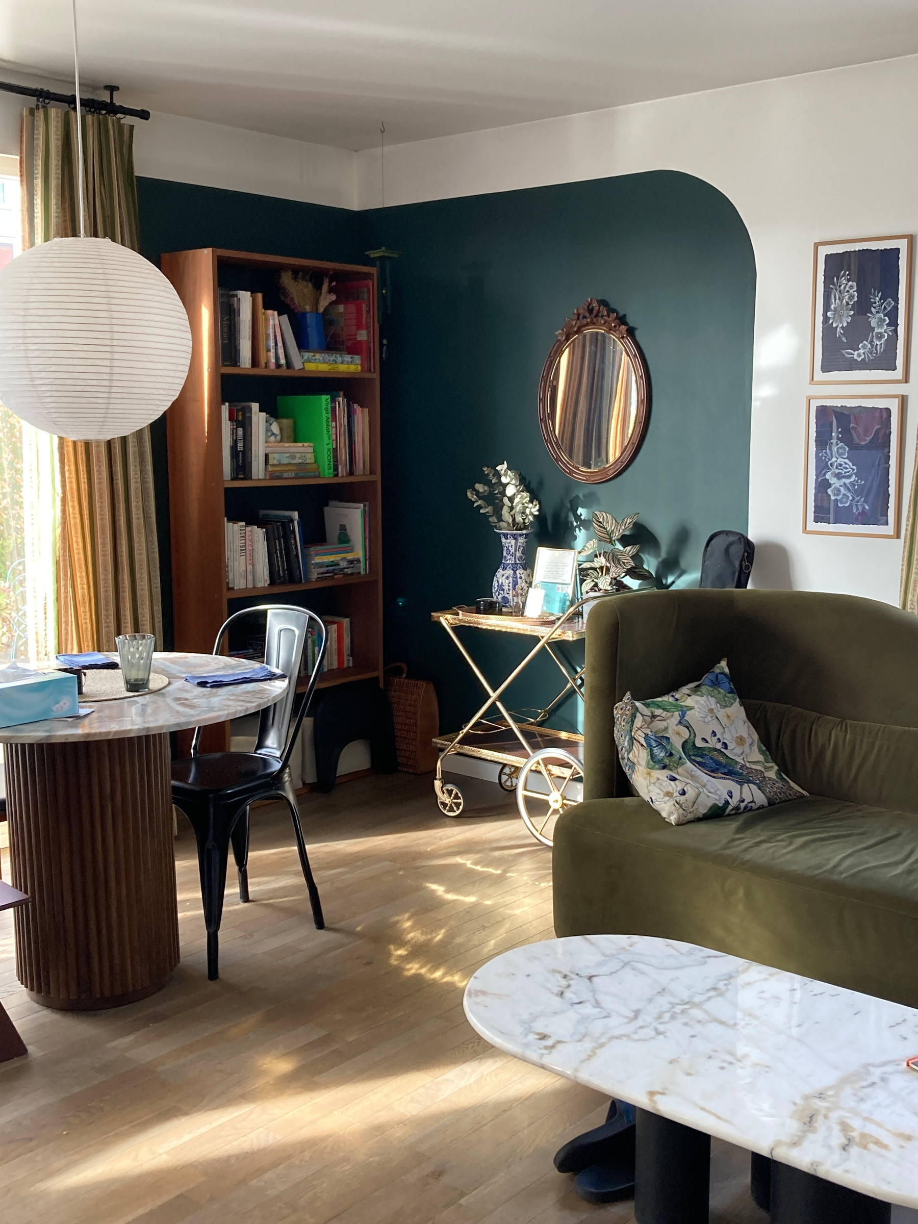 A cozy living room features a marble coffee table, a green velvet sofa, a wooden bookshelf, and decorative elements like a round mirror and framed art.