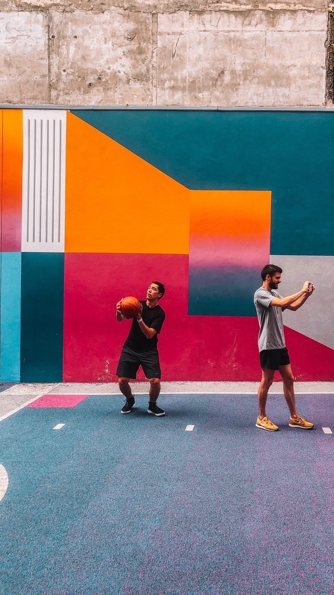 Two men play on a vibrant, colorful basketball court with geometric wall art in the background.