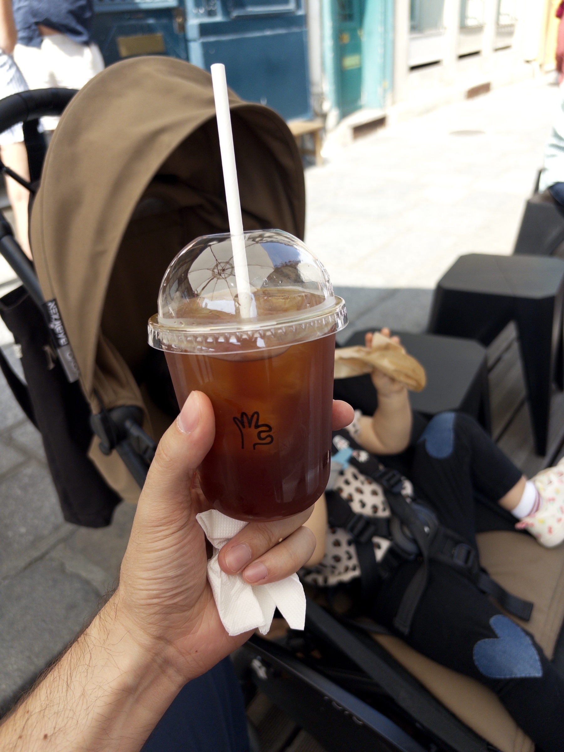A hand is holding a cup of iced coffee in front of a stroller, where a child is sitting and eating.