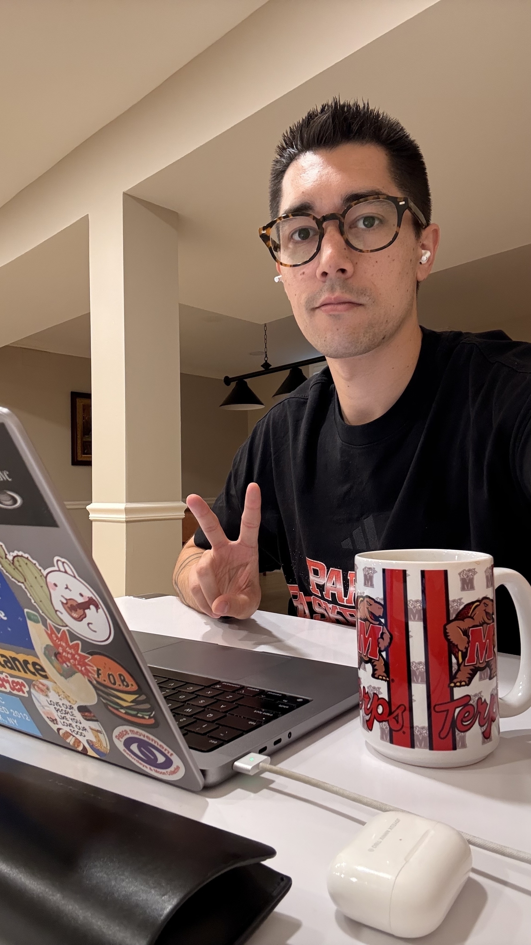 A person wearing glasses and a black T-shirt is seated at a table with a laptop and a Maryland-themed mug, making a peace sign.