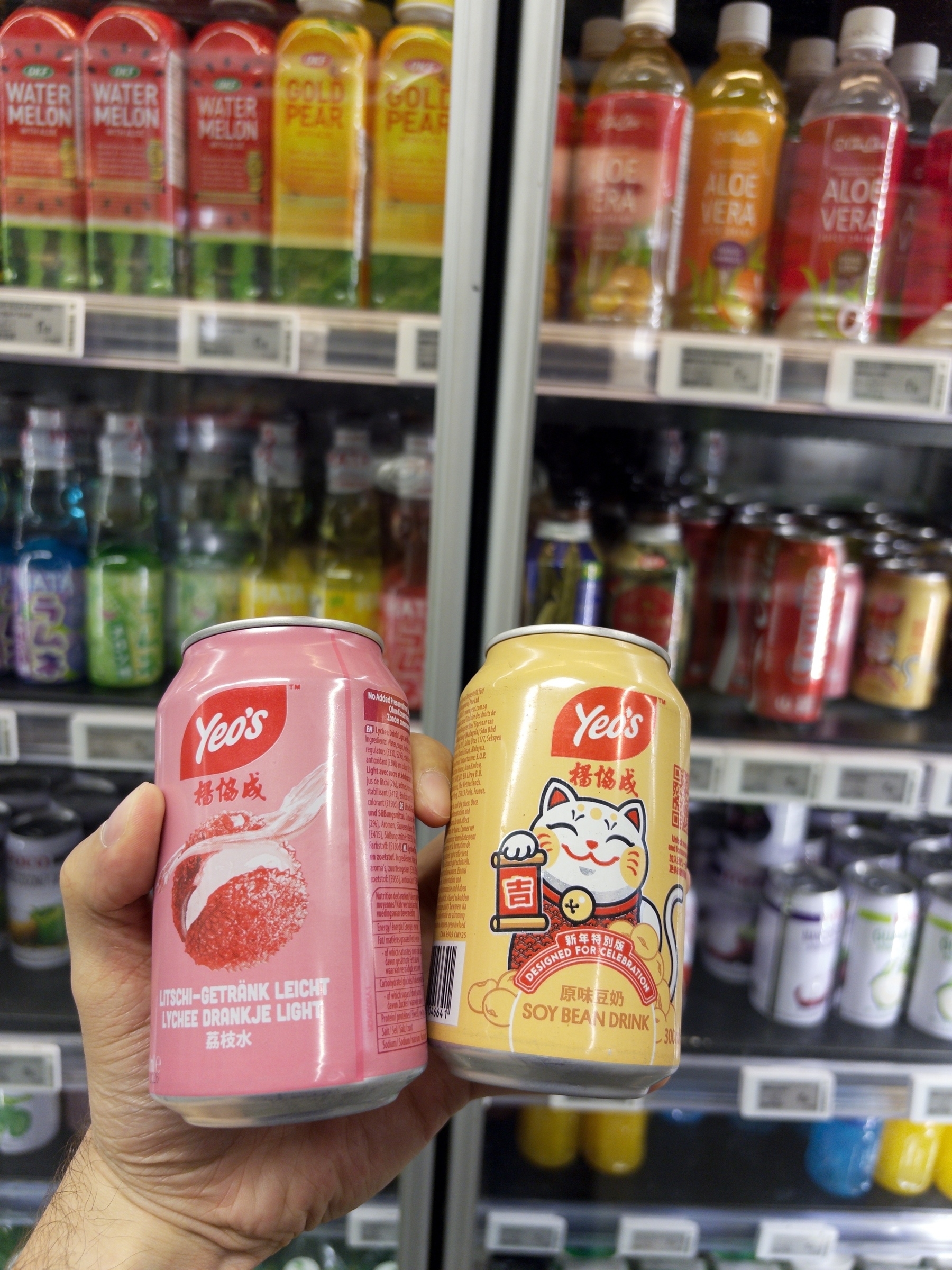 A person is holding two cans of Yeo's drinks, one lychee-flavored and the other soy bean, in front of a refrigerated display stocked with various beverages.