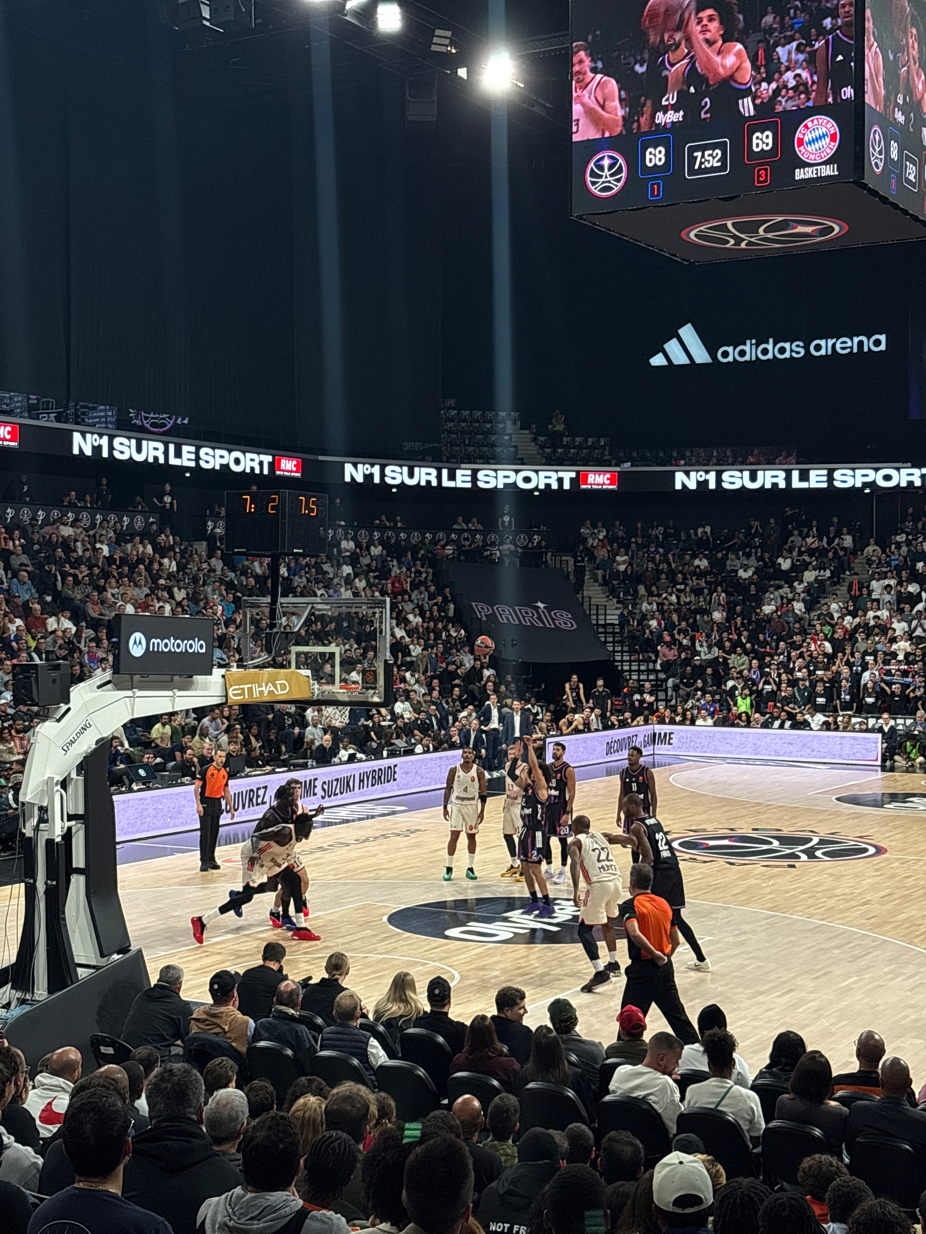 A basketball game is taking place in a packed arena, with players on the court and the score displayed on a large screen above.