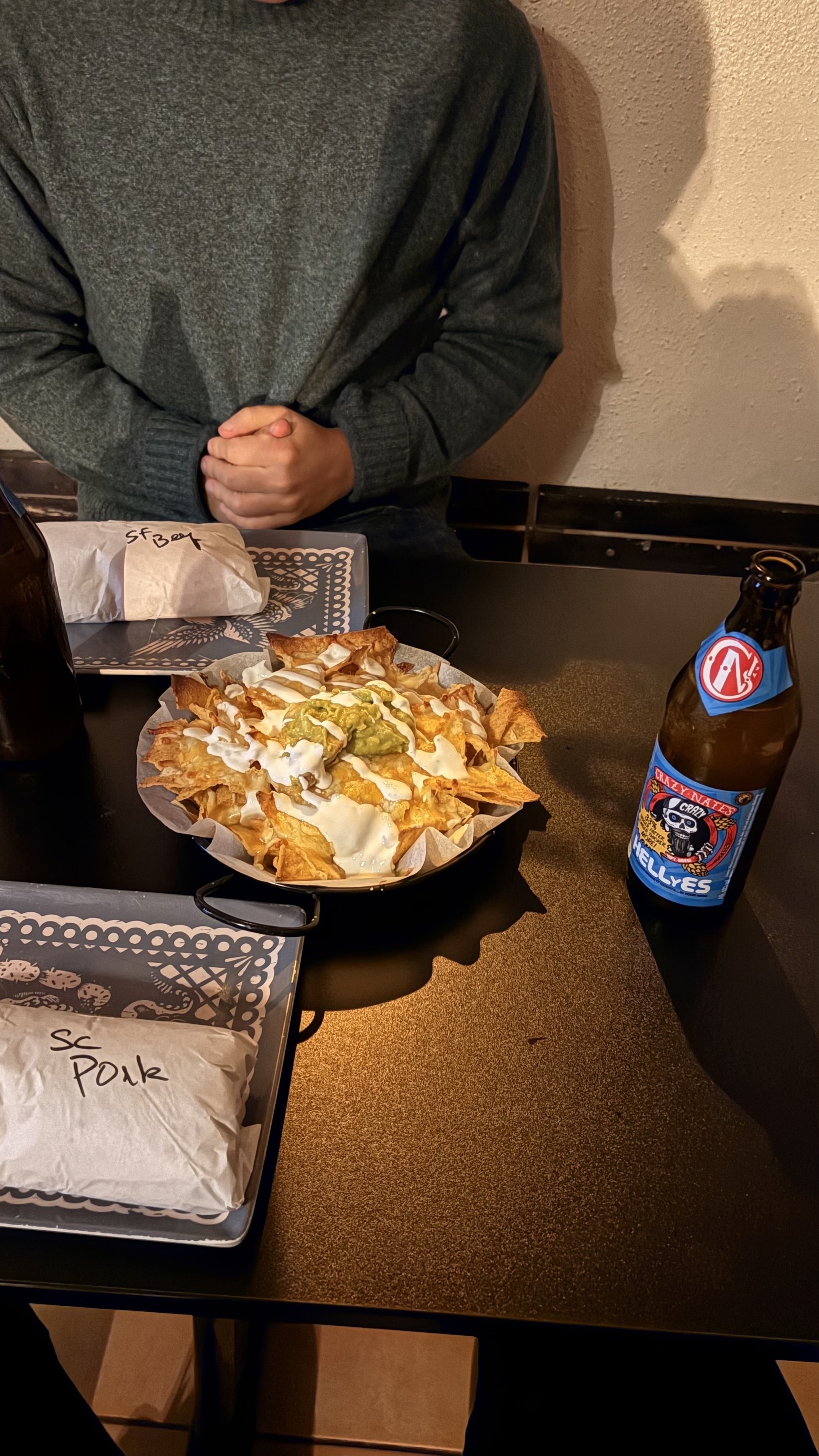 A person wearing a sweater is sitting at a table with a dish of nachos, two wrapped sandwiches, and a bottle of Blue Moon beer.