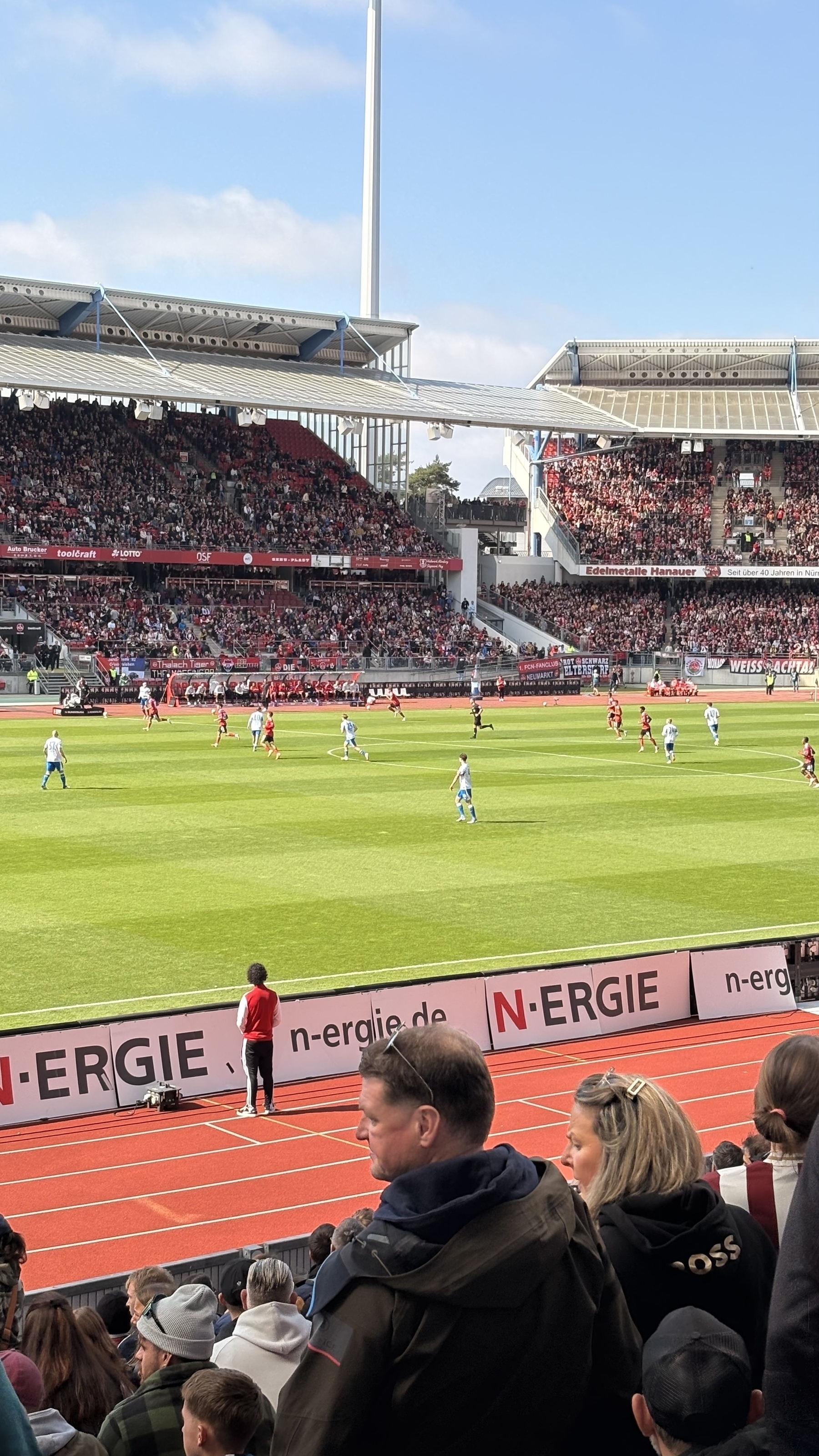 A lively soccer match takes place in a stadium filled with spectators under a clear blue sky.