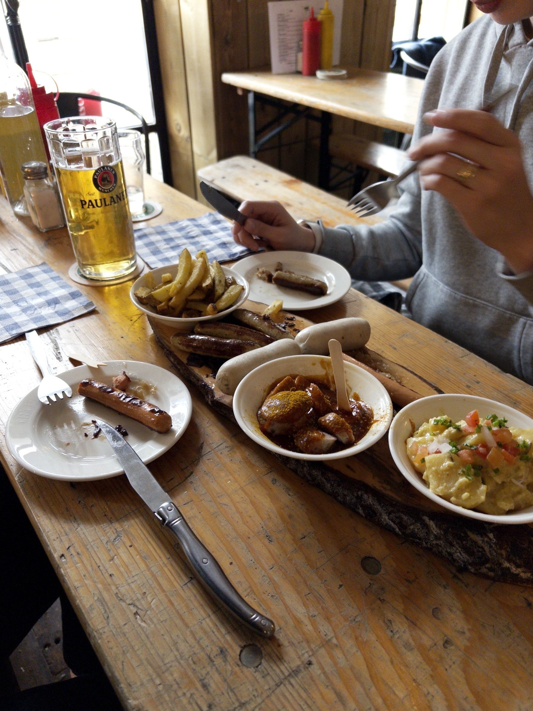 Two people are enjoying a meal with sausages, potatoes, and salad accompanied by a glass of beer.