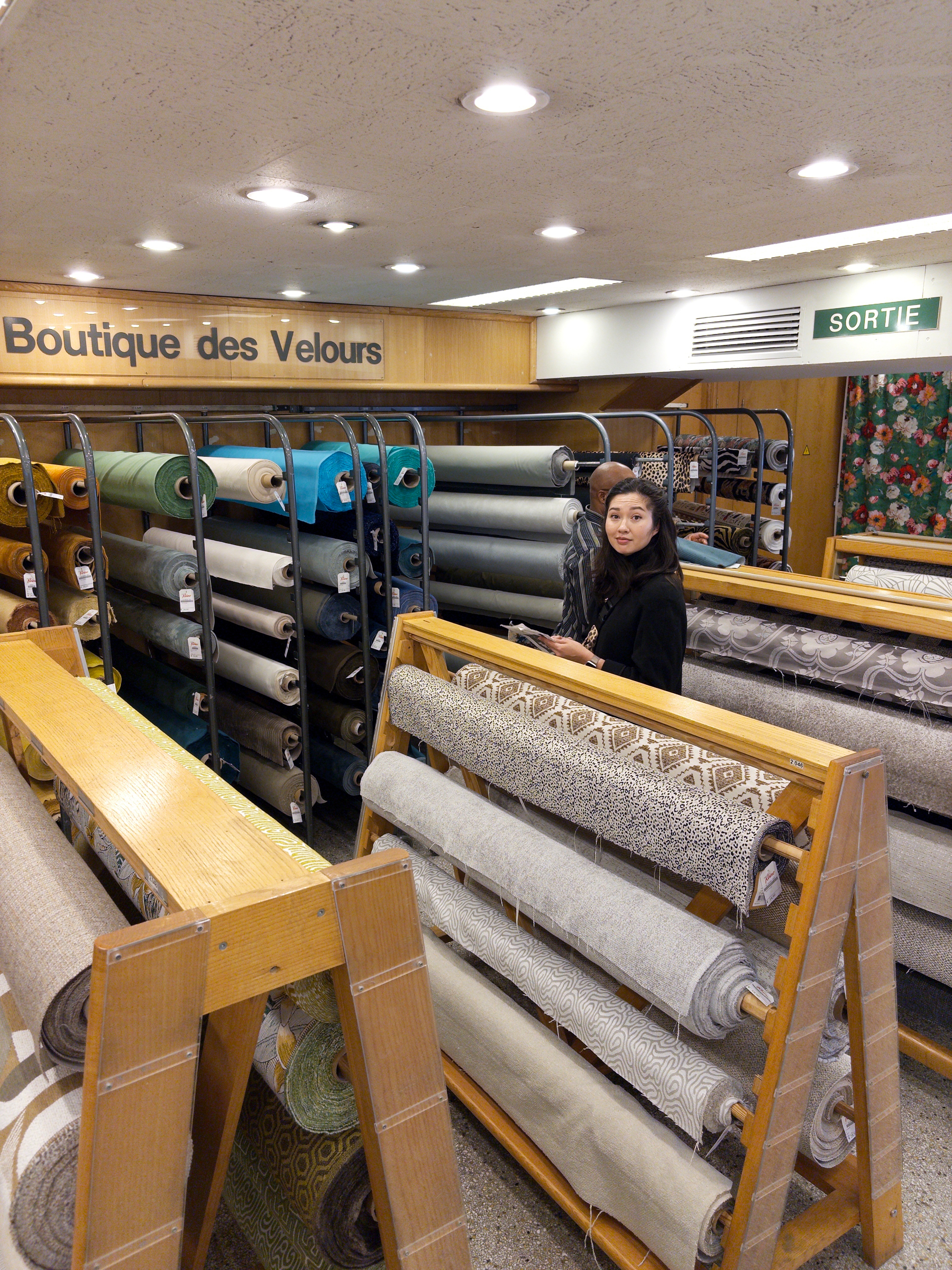 A person is browsing through rolls of fabric in a store labeled Boutique des Velours.