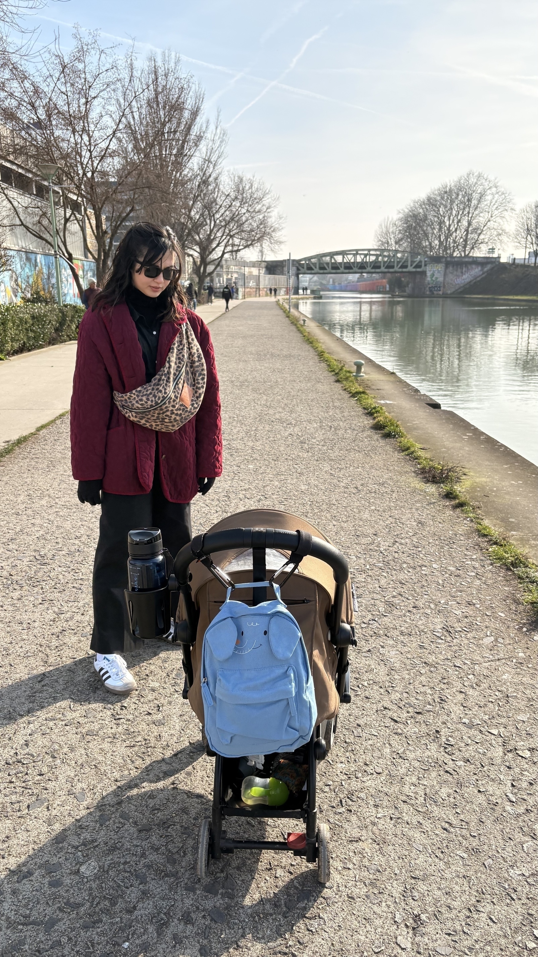 A woman wearing sunglasses stands next to a stroller with a blue bag attached, on a pathway beside a river.