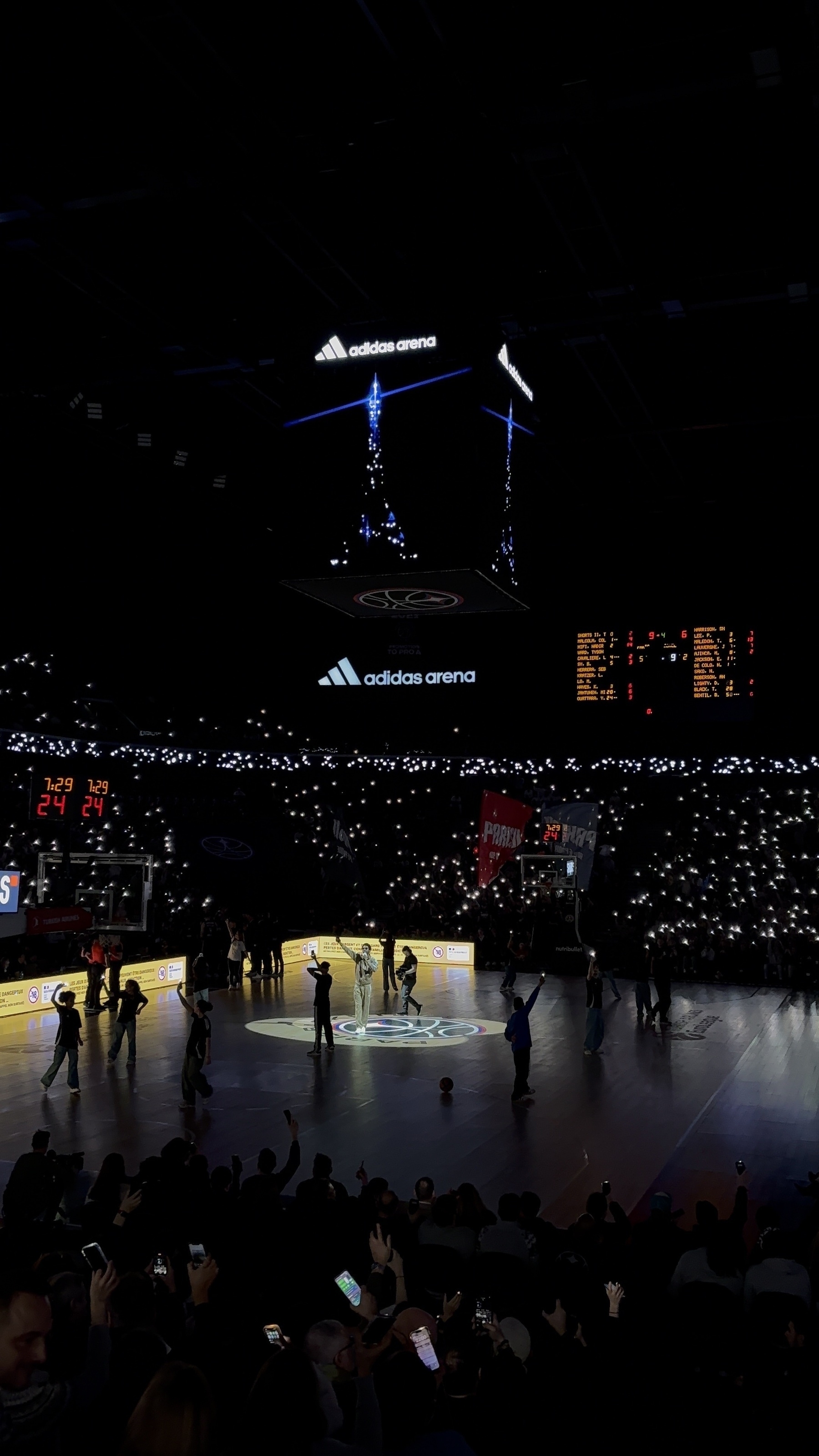 A basketball court with players and many spectators holding up lights in a dark arena.