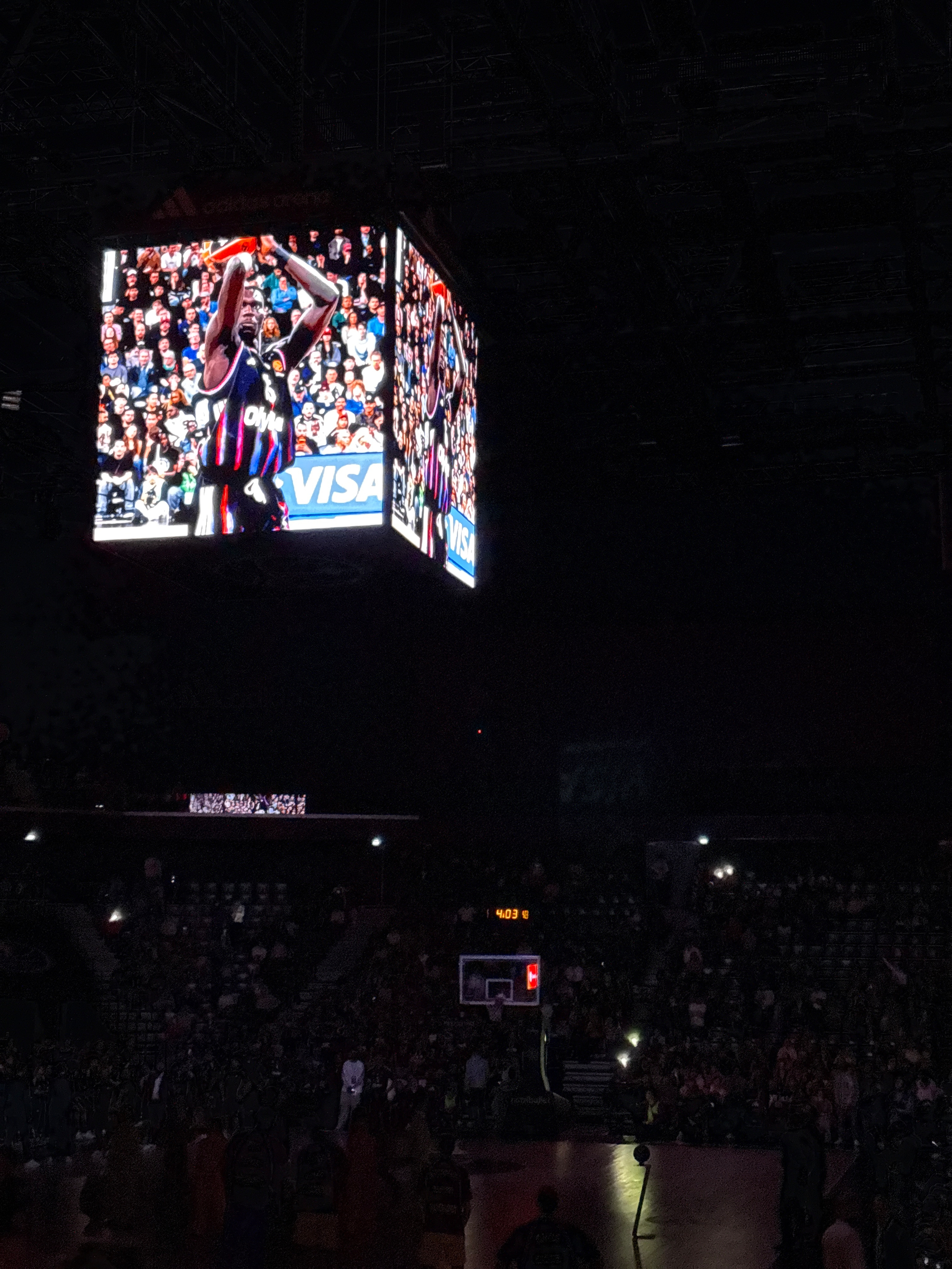 A basketball game is being played in a dimly lit arena with an overhead screen showing game highlights.