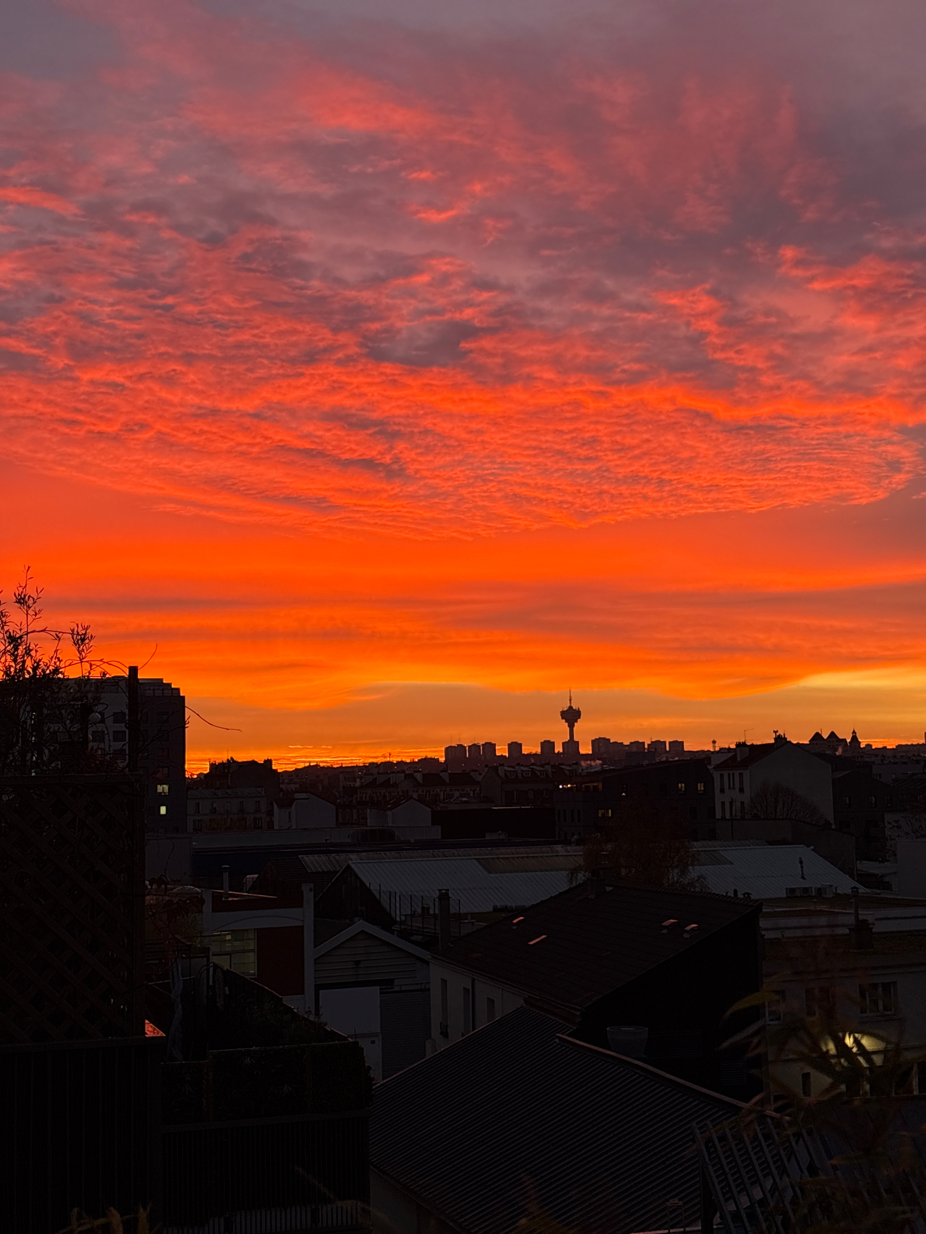 A vibrant sunrise with intense orange and red hues illuminates the skyline of a city, featuring various buildings and a tower silhouette in the distance.
